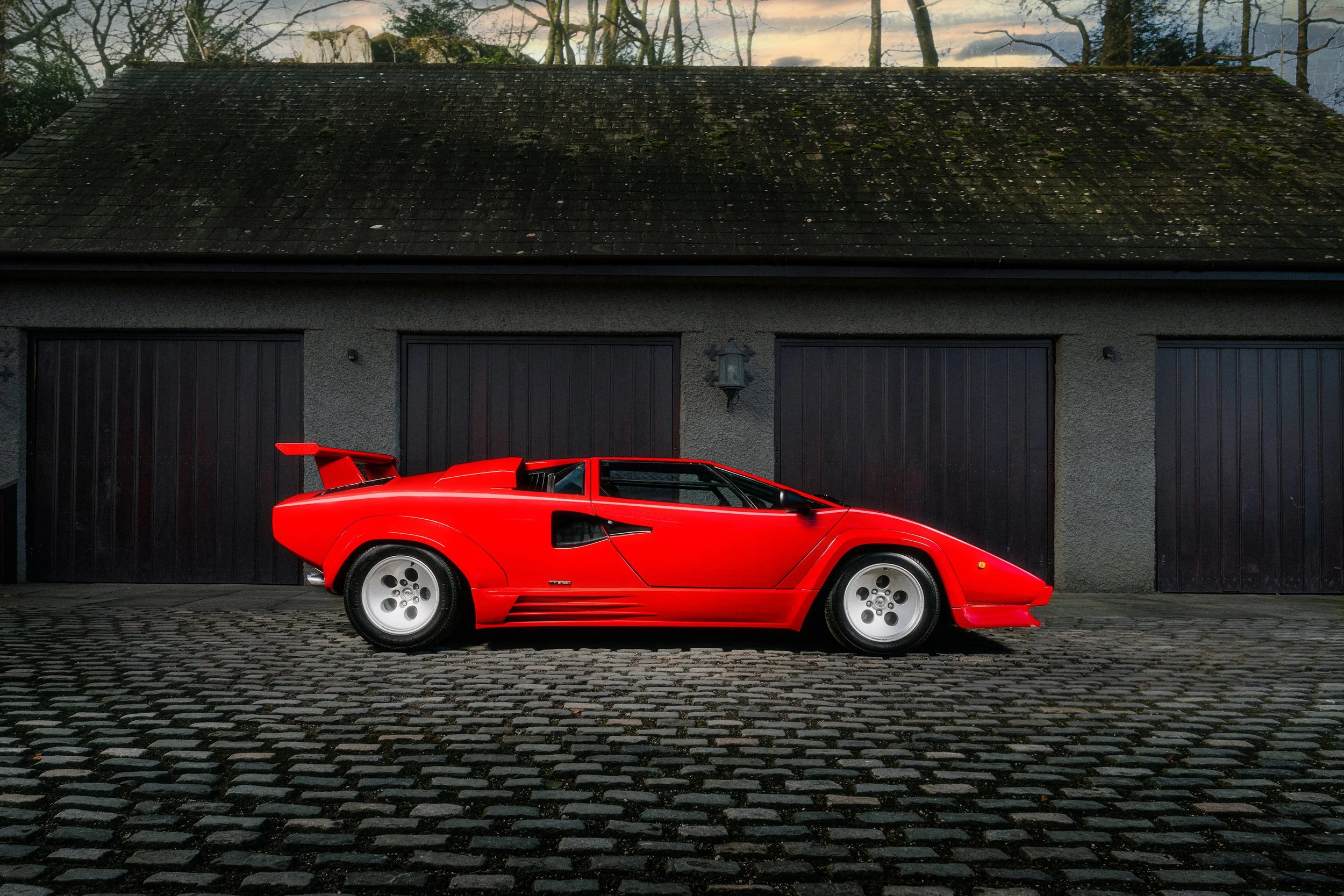A red Lamborghini Countach parked on a cobblestone driveway in front of a garage with dark doors, under a cloudy sky.