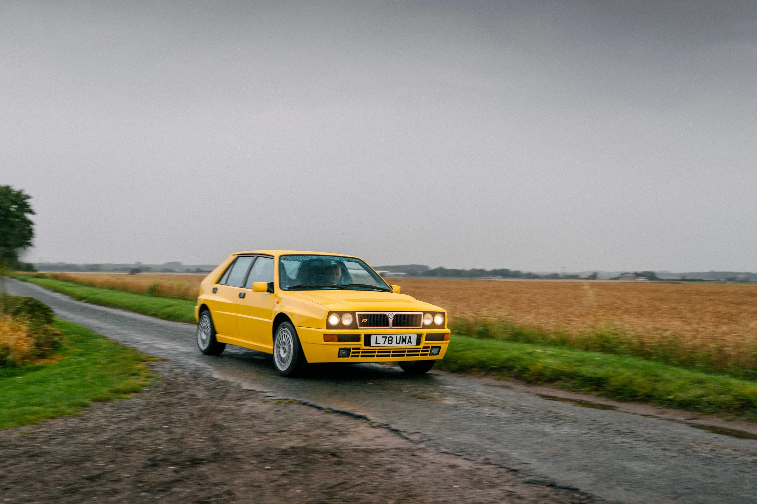 A yellow vintage hatchback car driving on a rural road with fields on either side under a gray overcast sky.