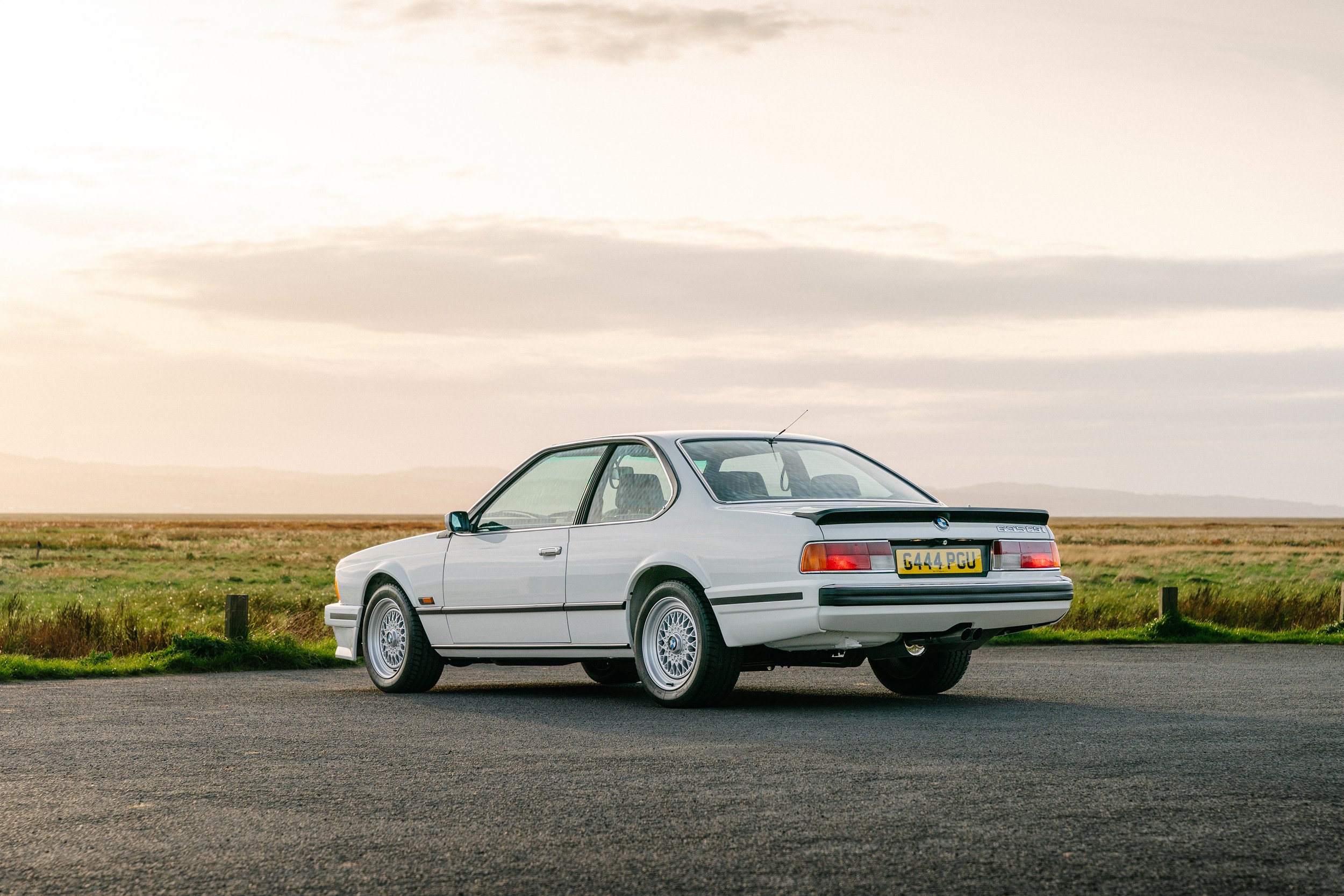 A white vintage BMW E24 6 Series parked on a road near a grassy field under a cloudy sky.