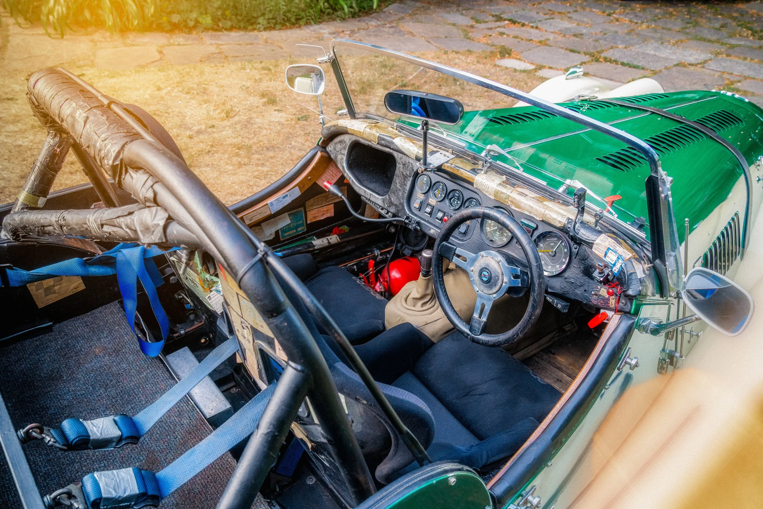 Interior of a vintage race car with a worn steering wheel, dashboard with gauges, racing seats with harnesses, and a roll cage.
