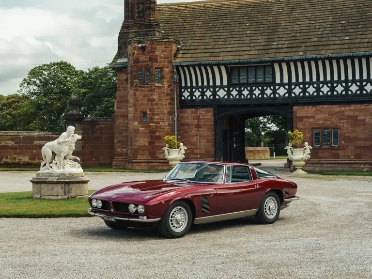 A vintage red sports car parked in front of a historic building with a brick and timber facade, stone statues, and large potted plants.