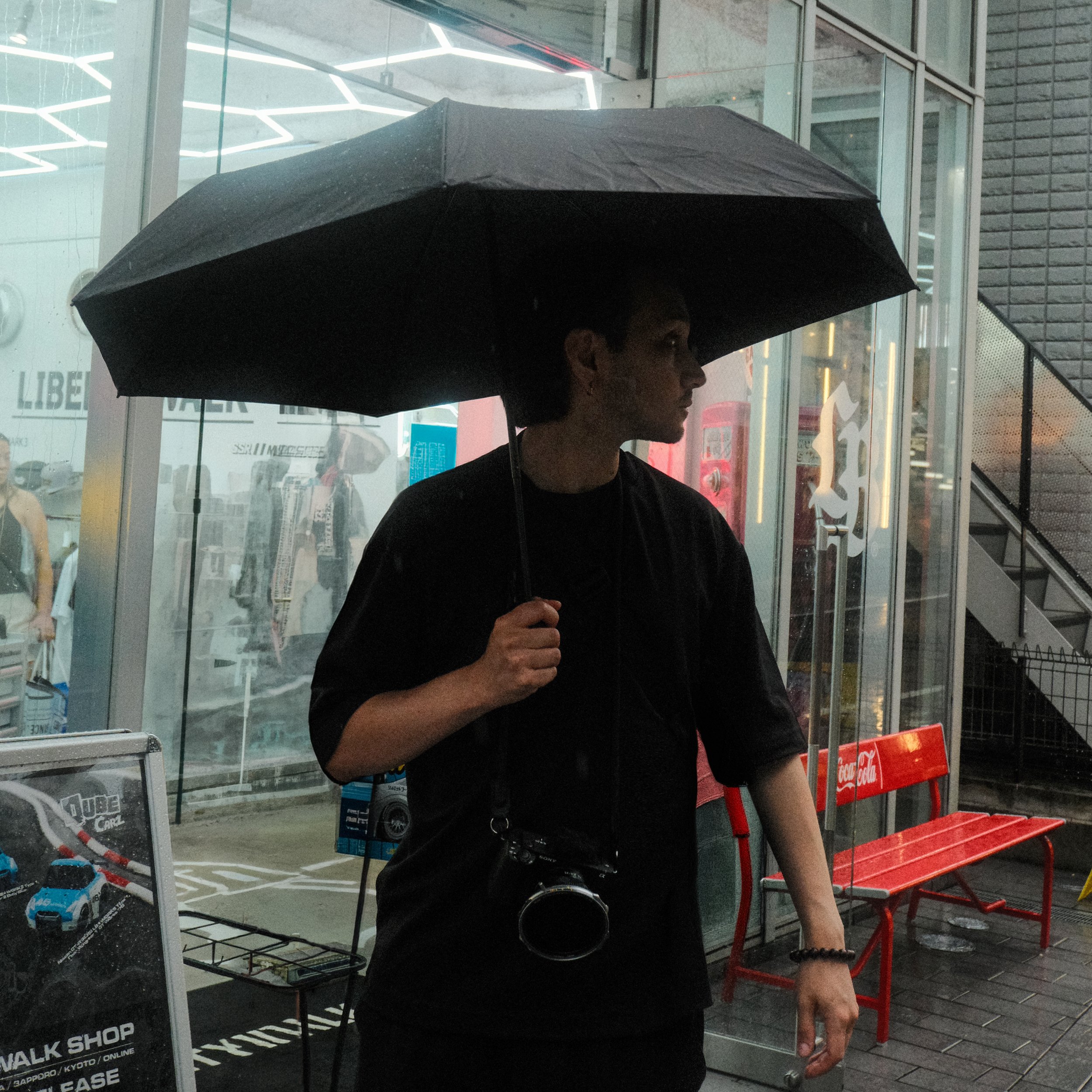 A young man holding an umbrella standing outside a building on a rainy day. The building has glass windows and a staircase, with red benches nearby.