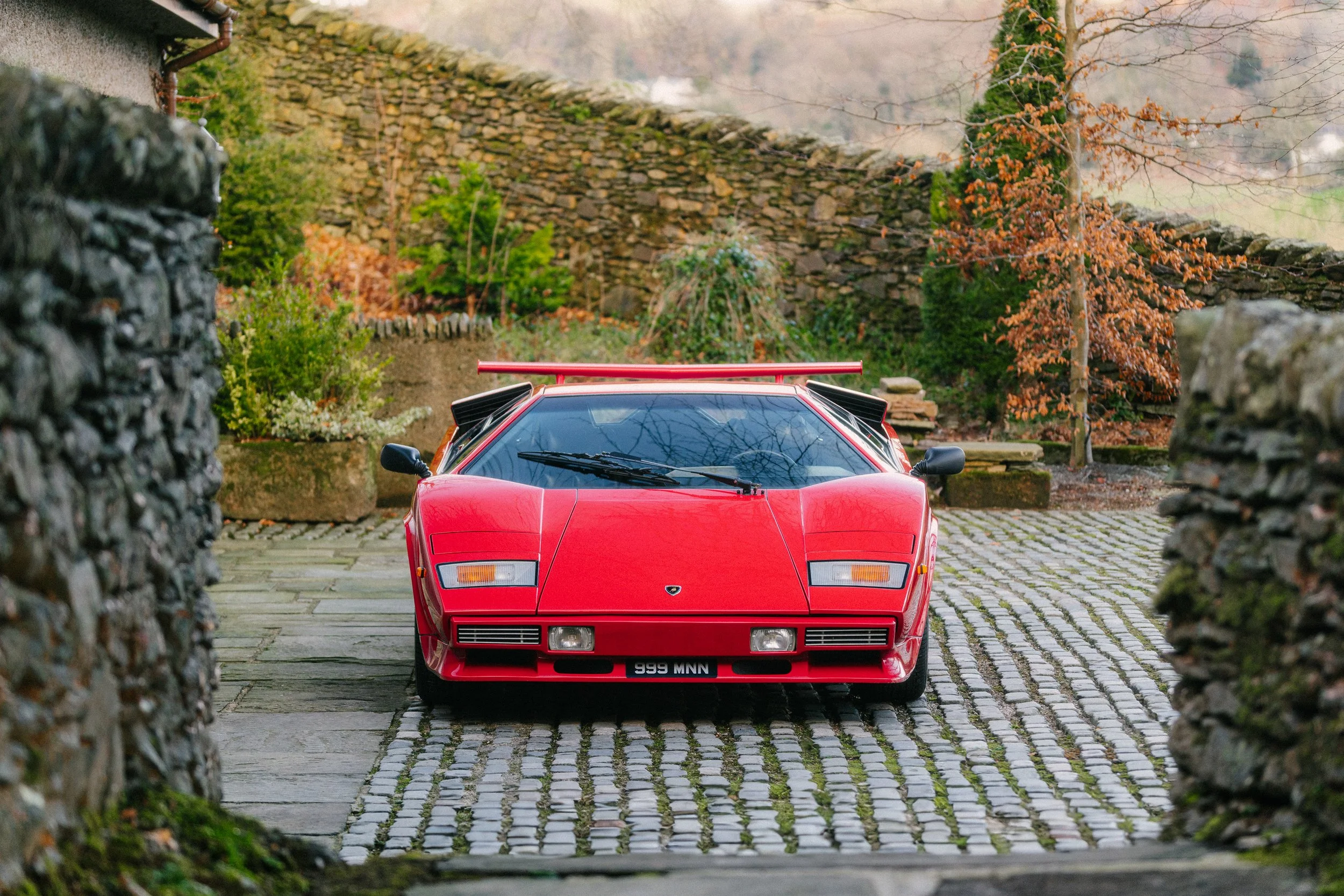 A red Lamborghini Countach sports car parked between stone walls with a cobblestone driveway in a garden setting.