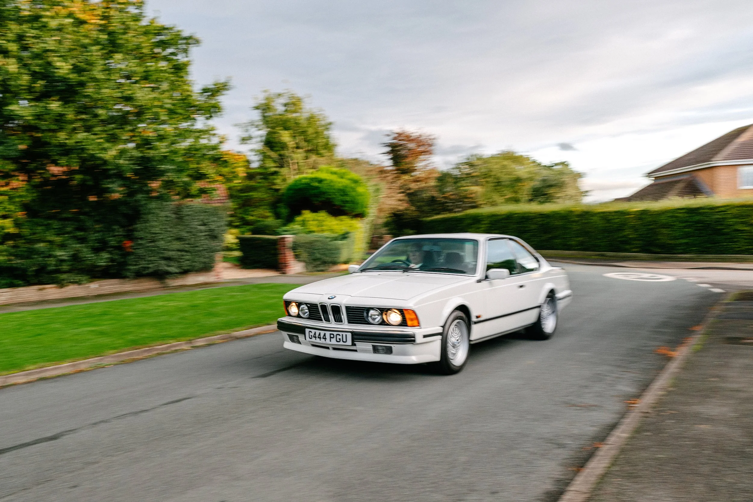 A white vintage BMW car driving on a residential street with greenery and houses in the background.