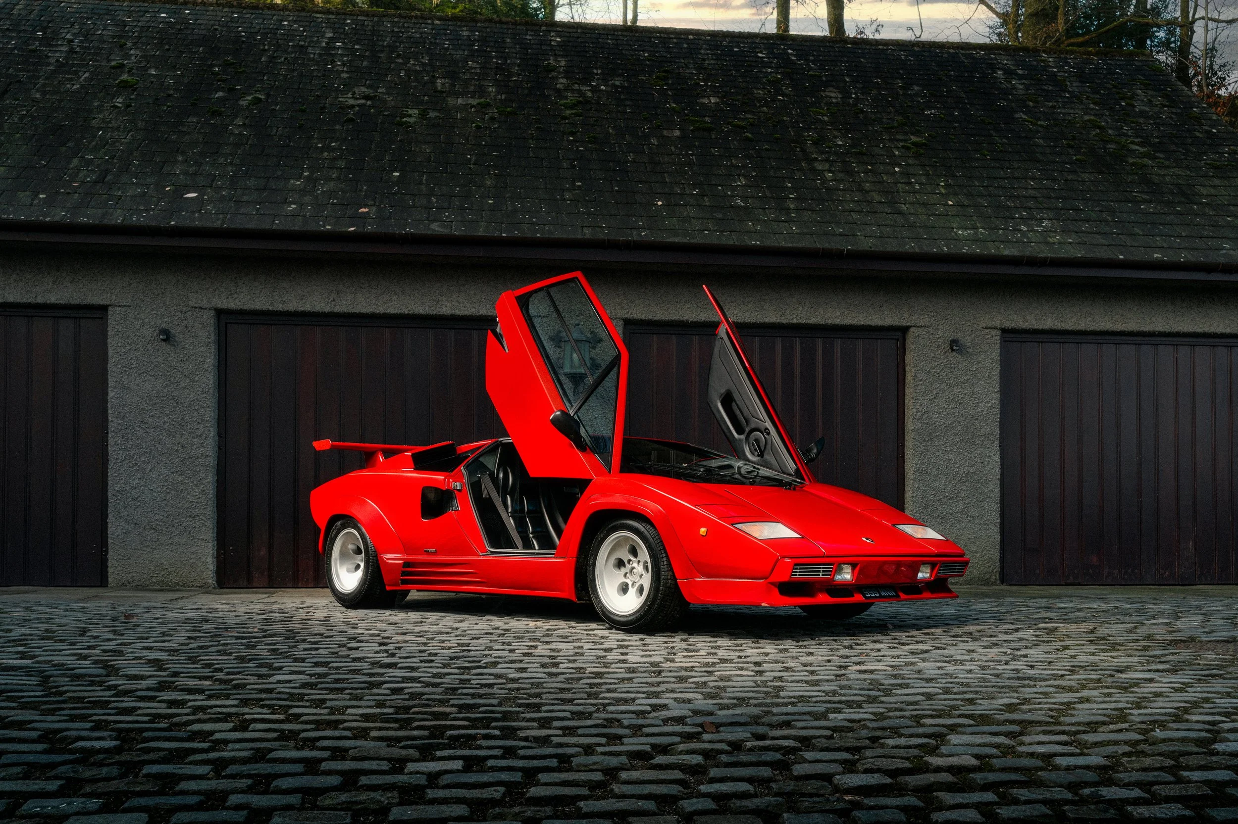 A classic red Lamborghini Countach with the driver's side door open, parked on a cobblestone driveway in front of a garage with dark wood doors.