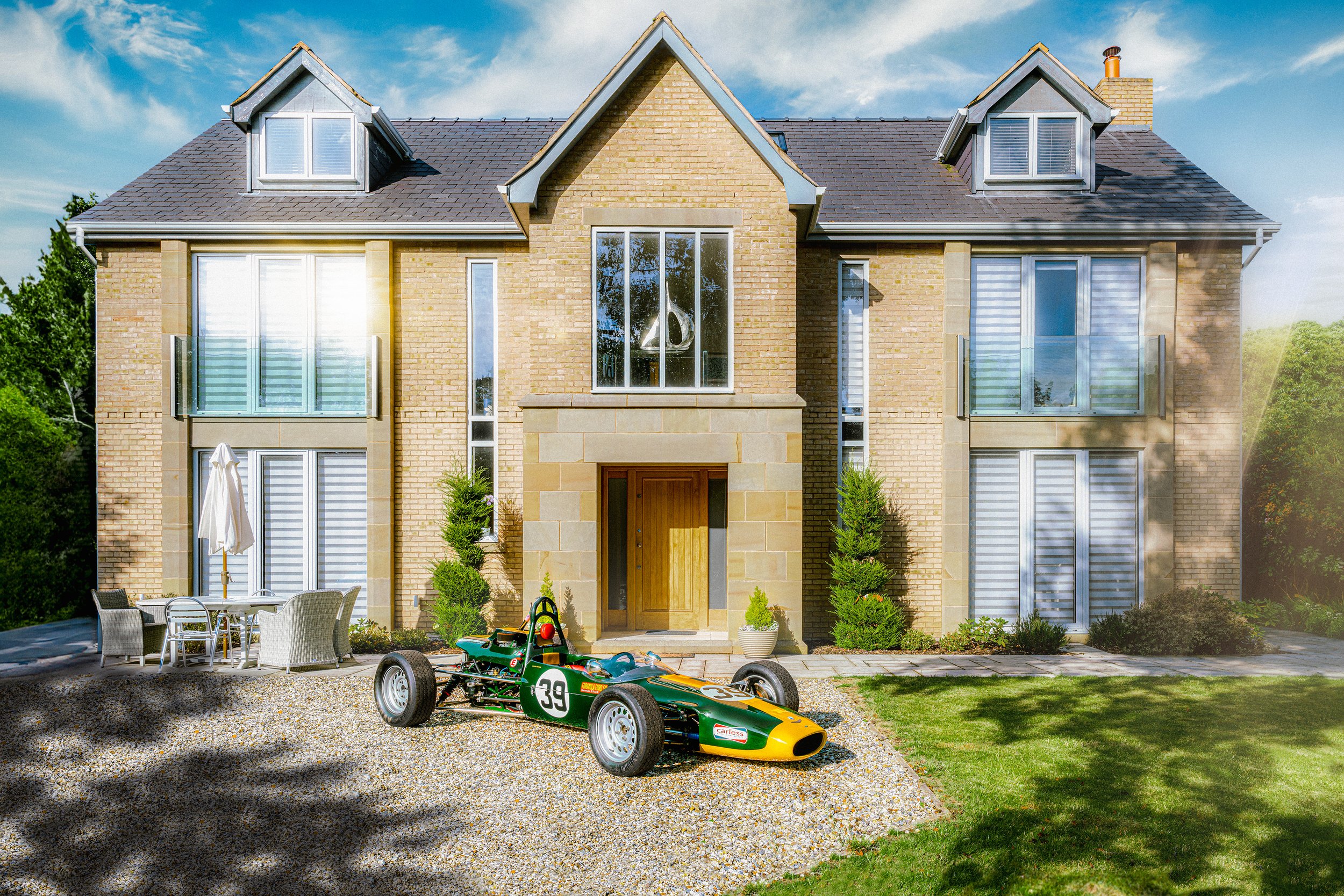 A modern two-story house with brick and stone exterior, large windows, a small front garden with bushes, a gravel driveway, a green vintage race car with the number 39, and outdoor seating with an umbrella. The sky is partly cloudy and trees line the