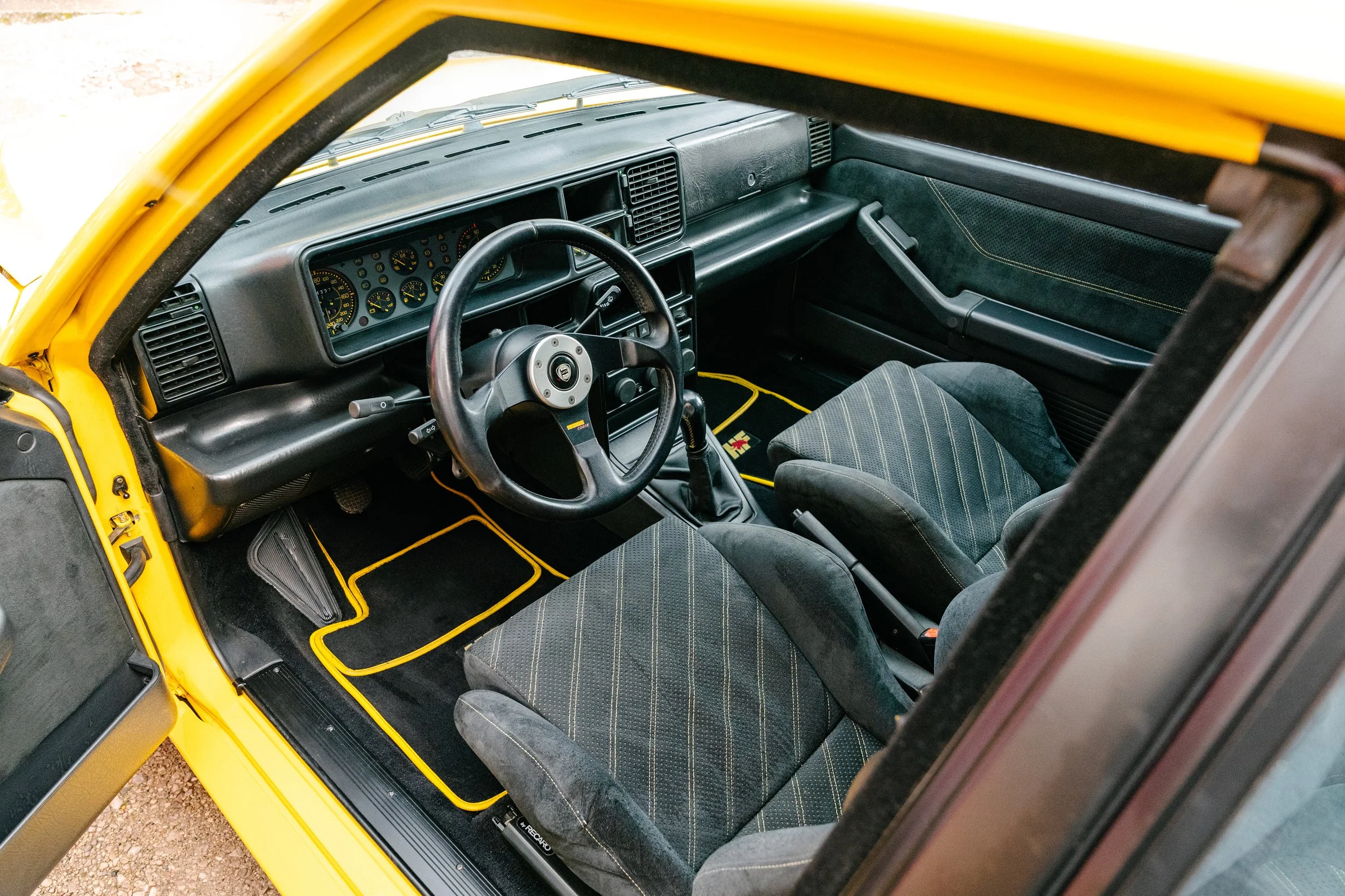 Inside view of a vintage yellow car showing the dashboard, steering wheel, and two cloth seats with yellow piping.