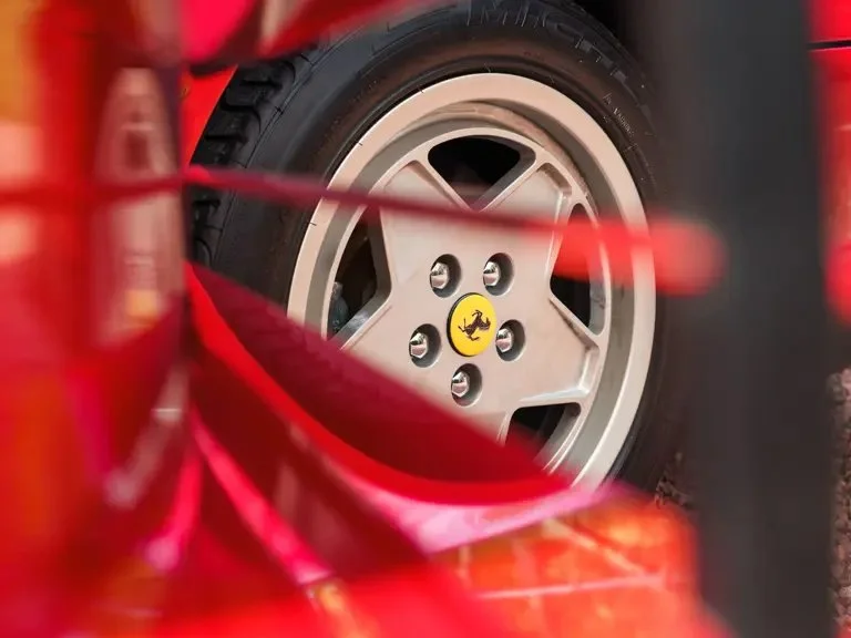 Close-up of a silver Ferrari wheel with a yellow Ferrari logo in the center, viewed through a red wire fence.