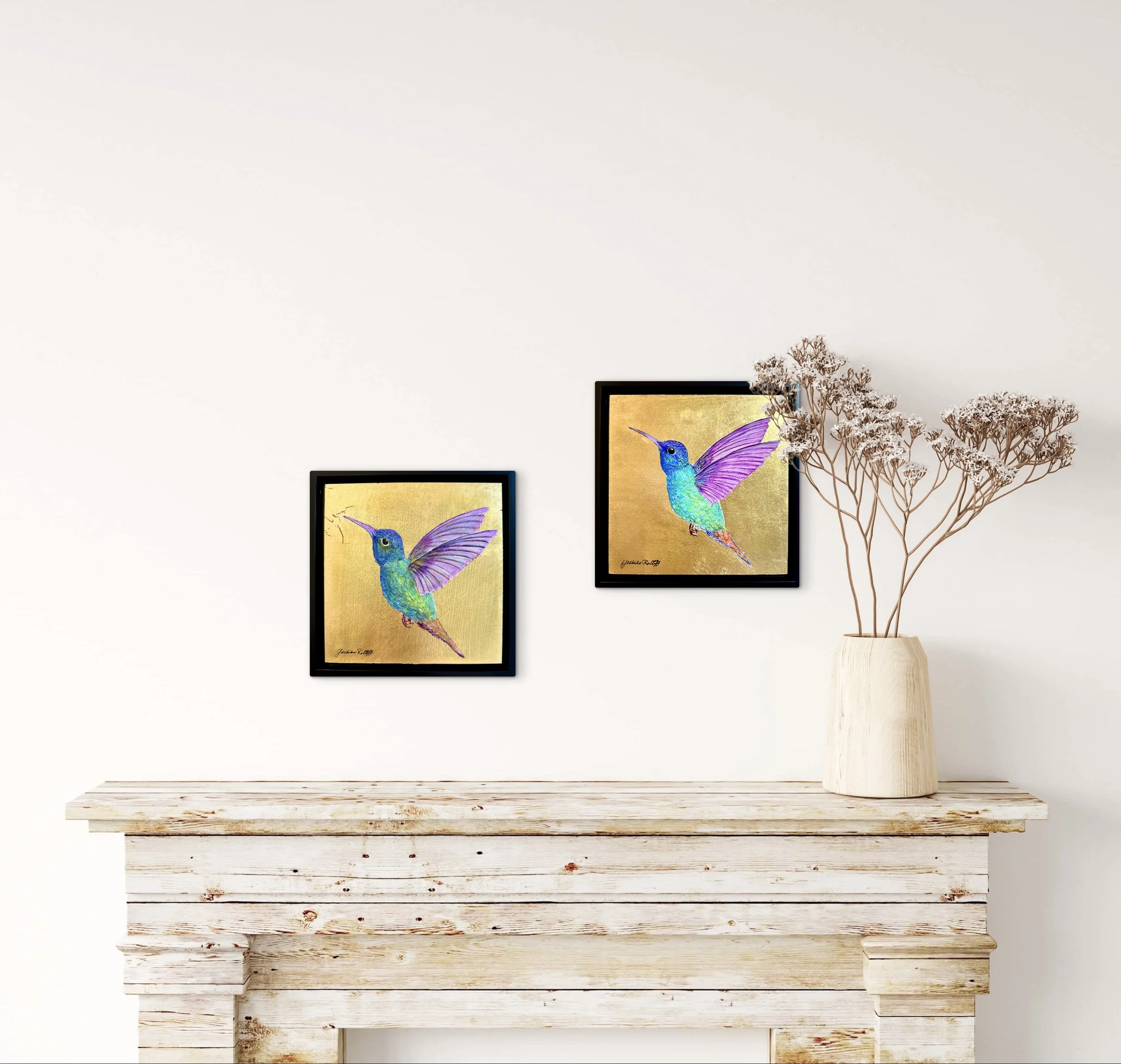 Decorative brown dried flowers in a beige vase on a white wooden table, with two framed original artworks of colorful hummingbirds hanging on a plain white wall in the background.