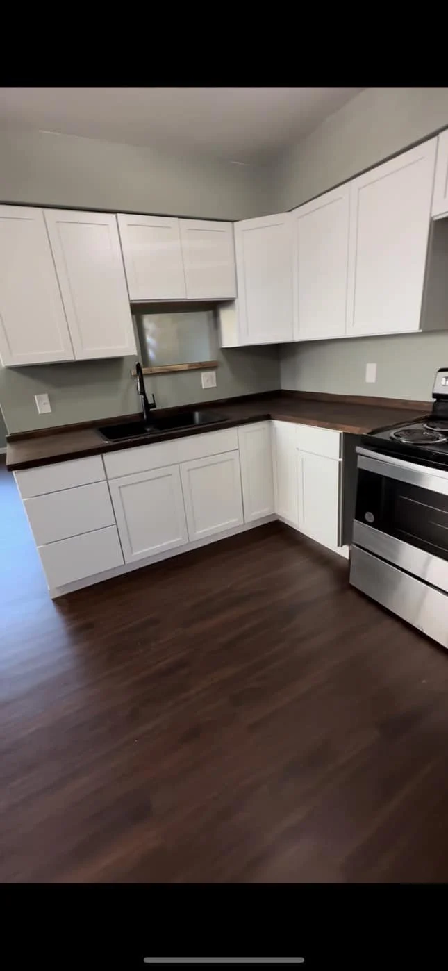 Modern kitchen with white cabinets, brown countertops, dark wood flooring, stainless steel oven, and a black sink.