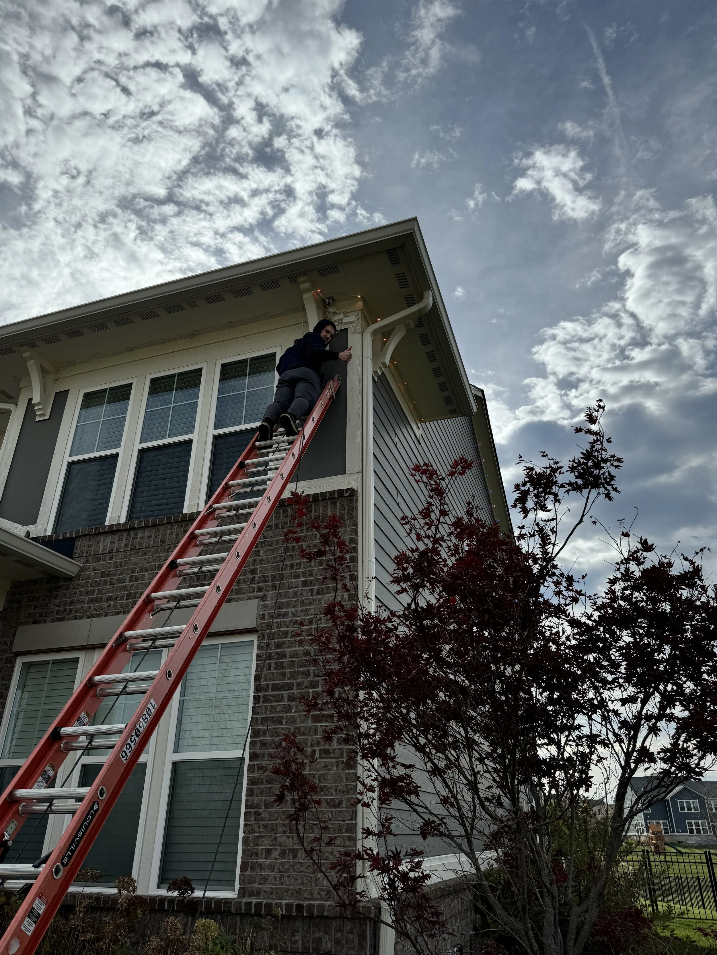 Person on a ladder hanging holiday lights on a two-story house with a brick facade and large windows. A tree is in the foreground, and the sky is partly cloudy.