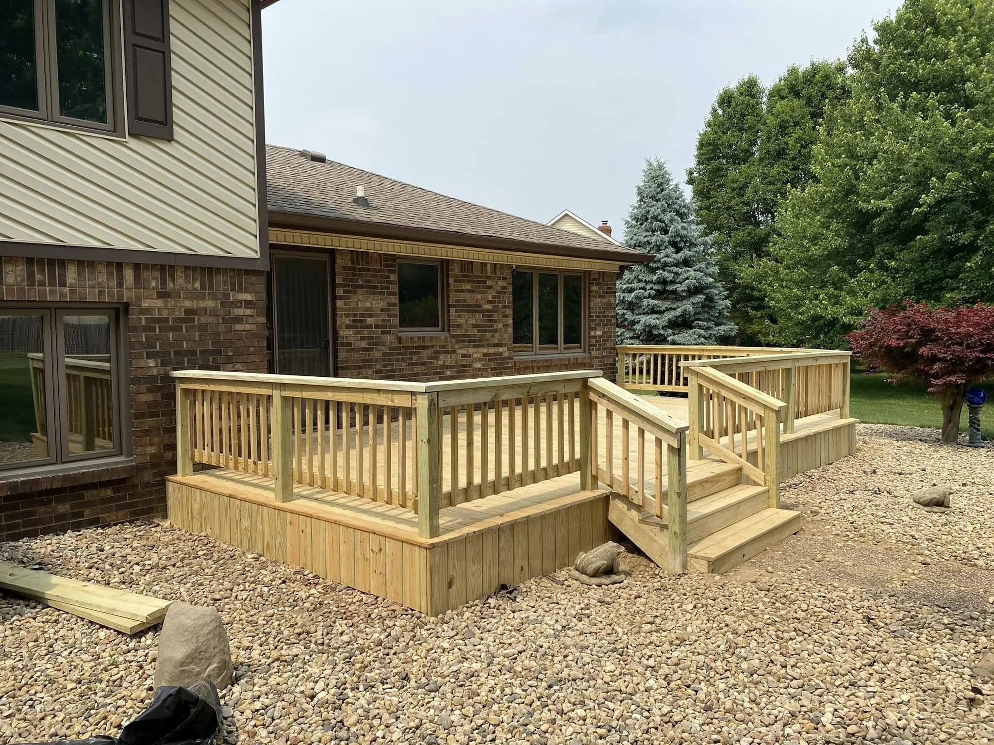 Newly constructed wooden deck with railings attached to a brick house, surrounded by a pebble-filled yard and trees in the background.