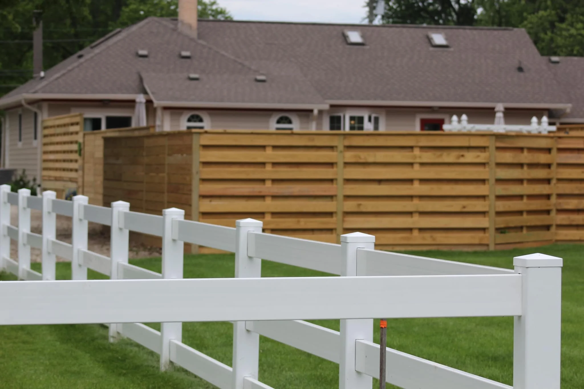 A white picket fence in front of a wooden privacy fence and a house with a gray roof and windows.