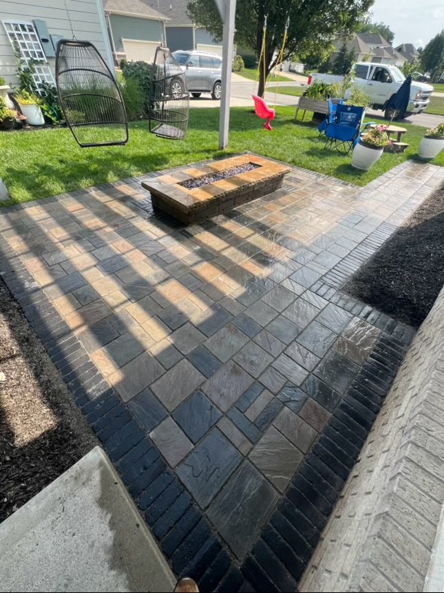Patio with a brick paver floor, featuring a rectangular fire pit, hanging chairs, and outdoor seating. Lawn and potted plants surround the area.