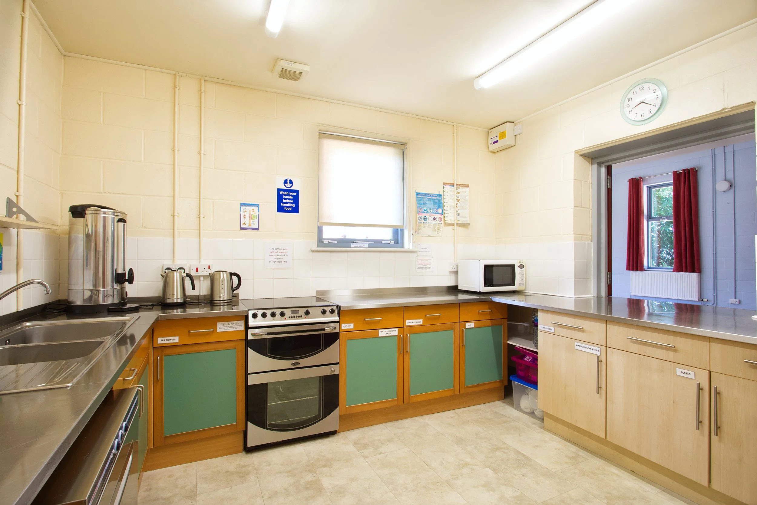 A clean kitchen with light-colored walls, green and wooden cabinets, a stove, microwave, and coffee-making appliances on the counter. There are windows with white and red curtains, a clock on the wall, and various notices and instructions posted.