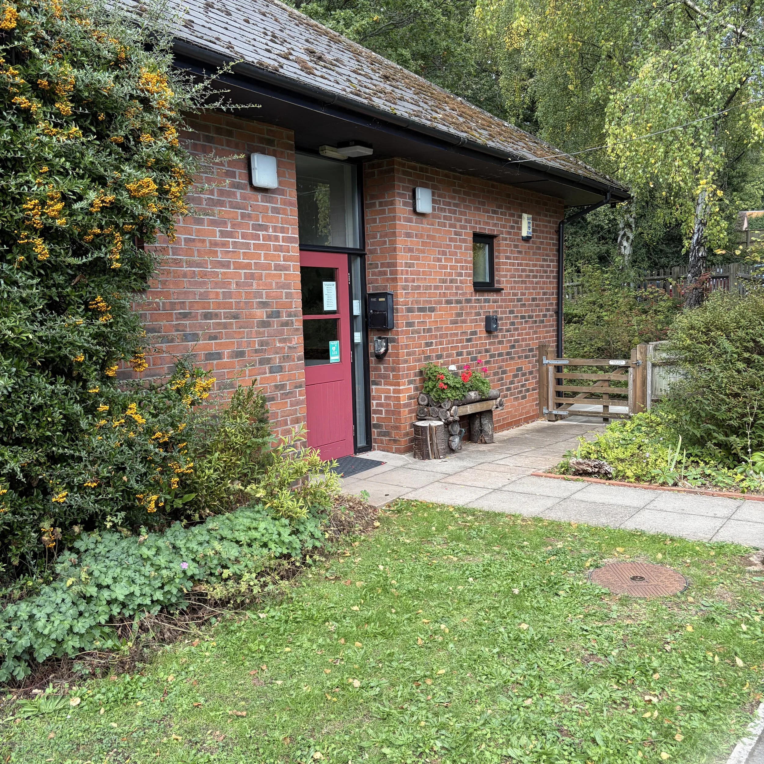 A brick building with large windows and a red door, surrounded by green plants and trees, with a small wooden fence and a paved sidewalk in front.