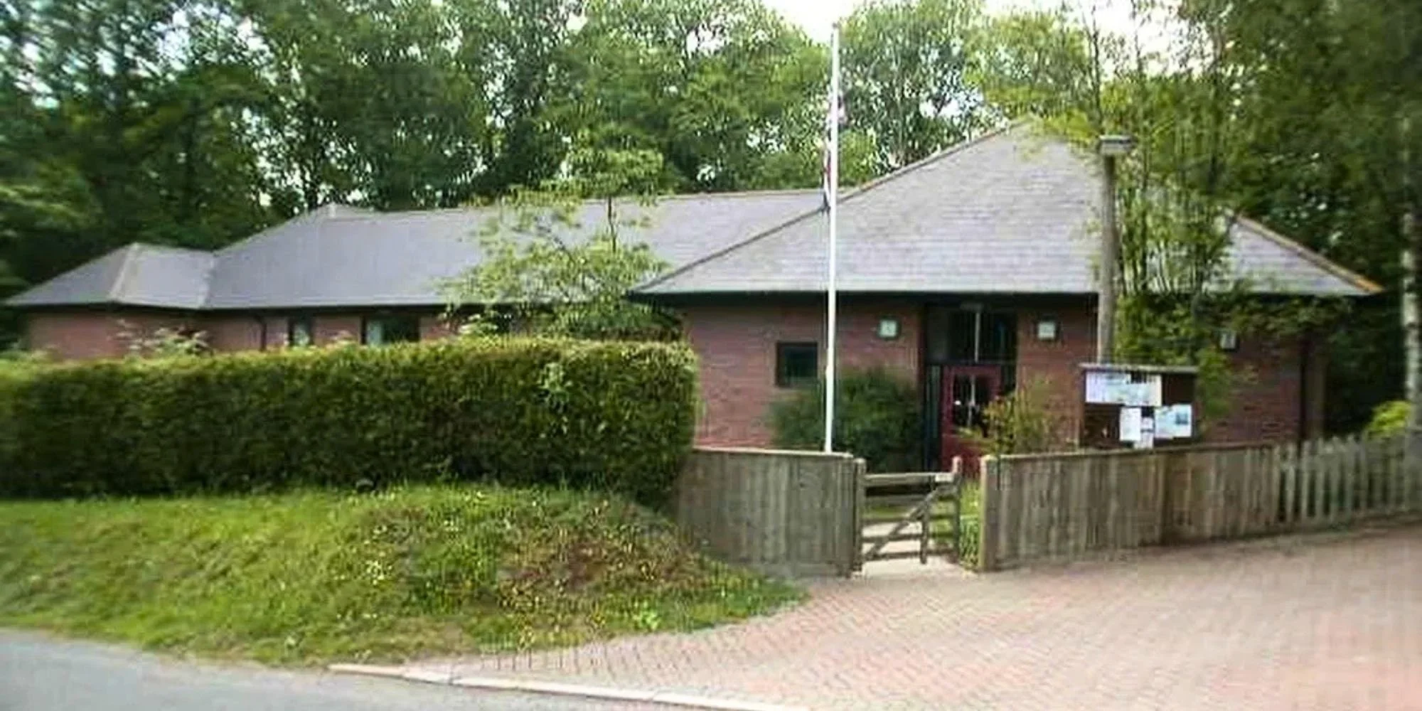 A red brick building with a gray pitched roof, surrounded by green trees and bushes. There is a wooden fence and a small gate at the front, with notices posted on a bulletin board near the entrance.