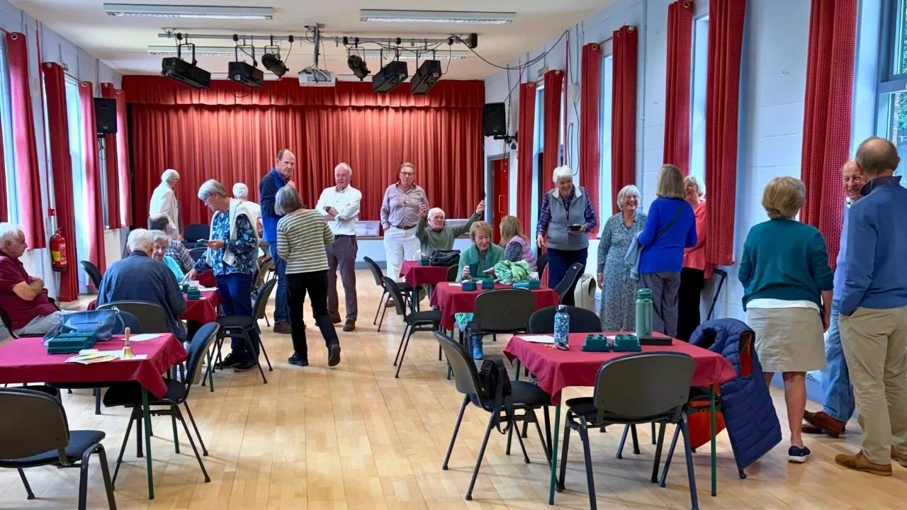 Group of older adults socializing in a community hall with red curtains and tables covered with red tablecloths.