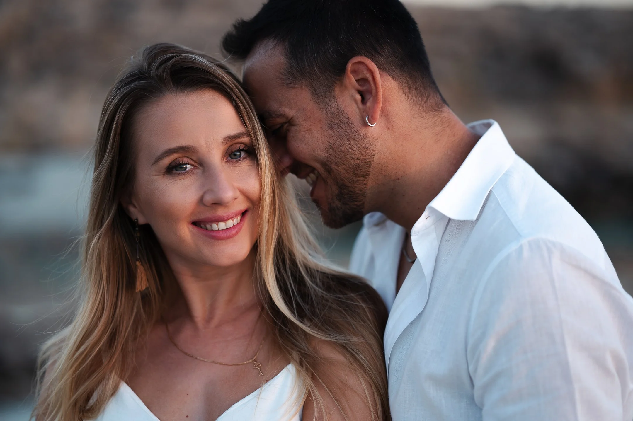 A smiling woman with long blonde hair and a man with short dark hair leaning their foreheads together outdoors at sunset.