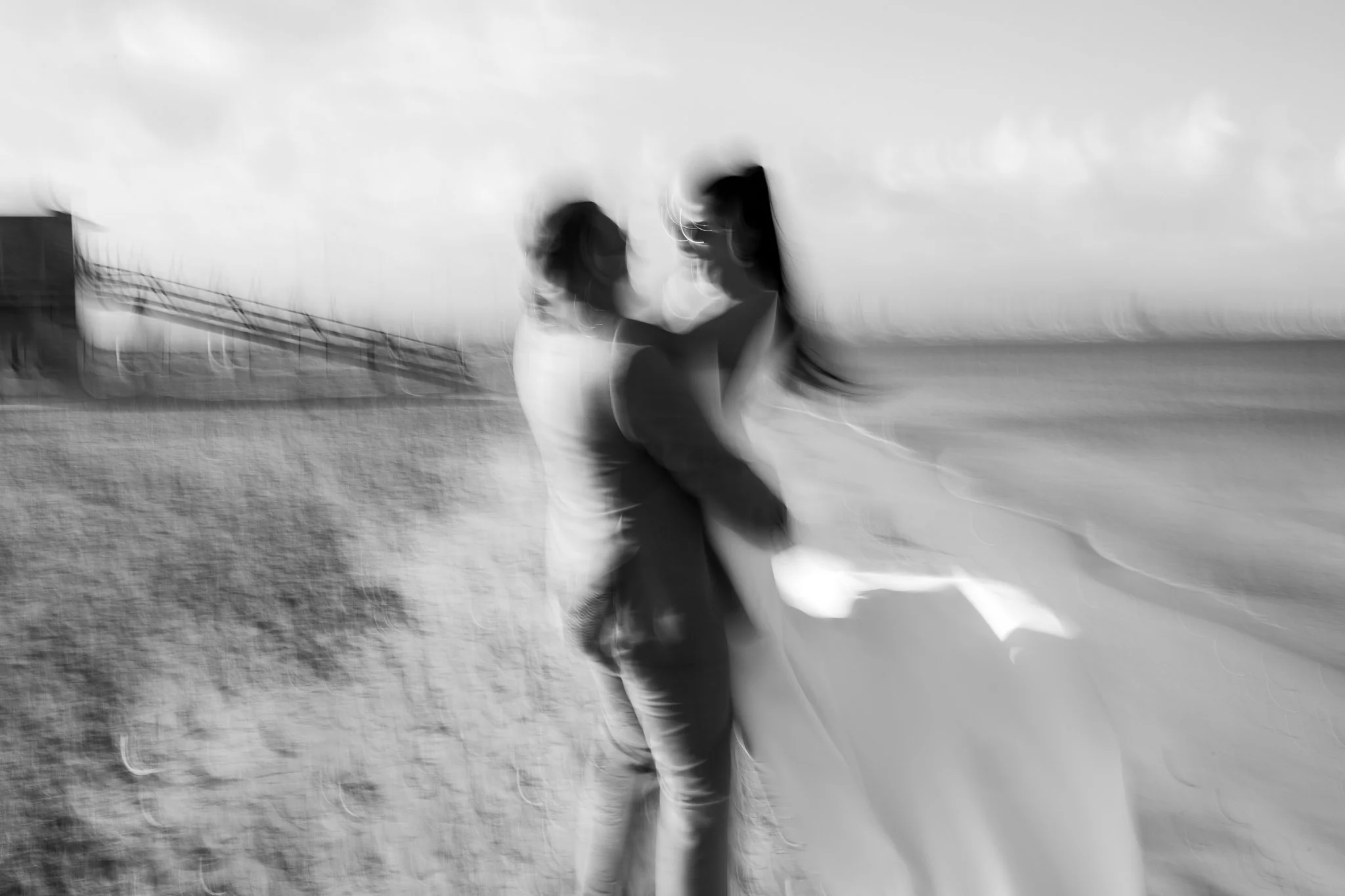 Blurry black and white photograph of two people standing outdoors near a water body, with a dock and cloudy sky in the background.