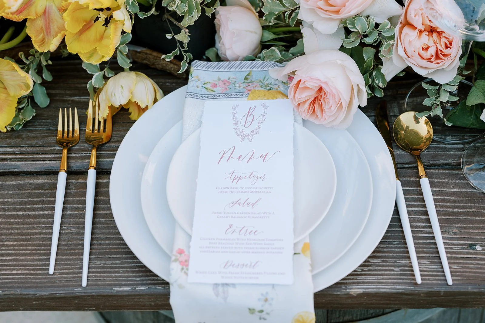 Elegant outdoor table setting with white plates, gold and white cutlery, a floral menu card, and pink peony flowers on a rustic wooden table.