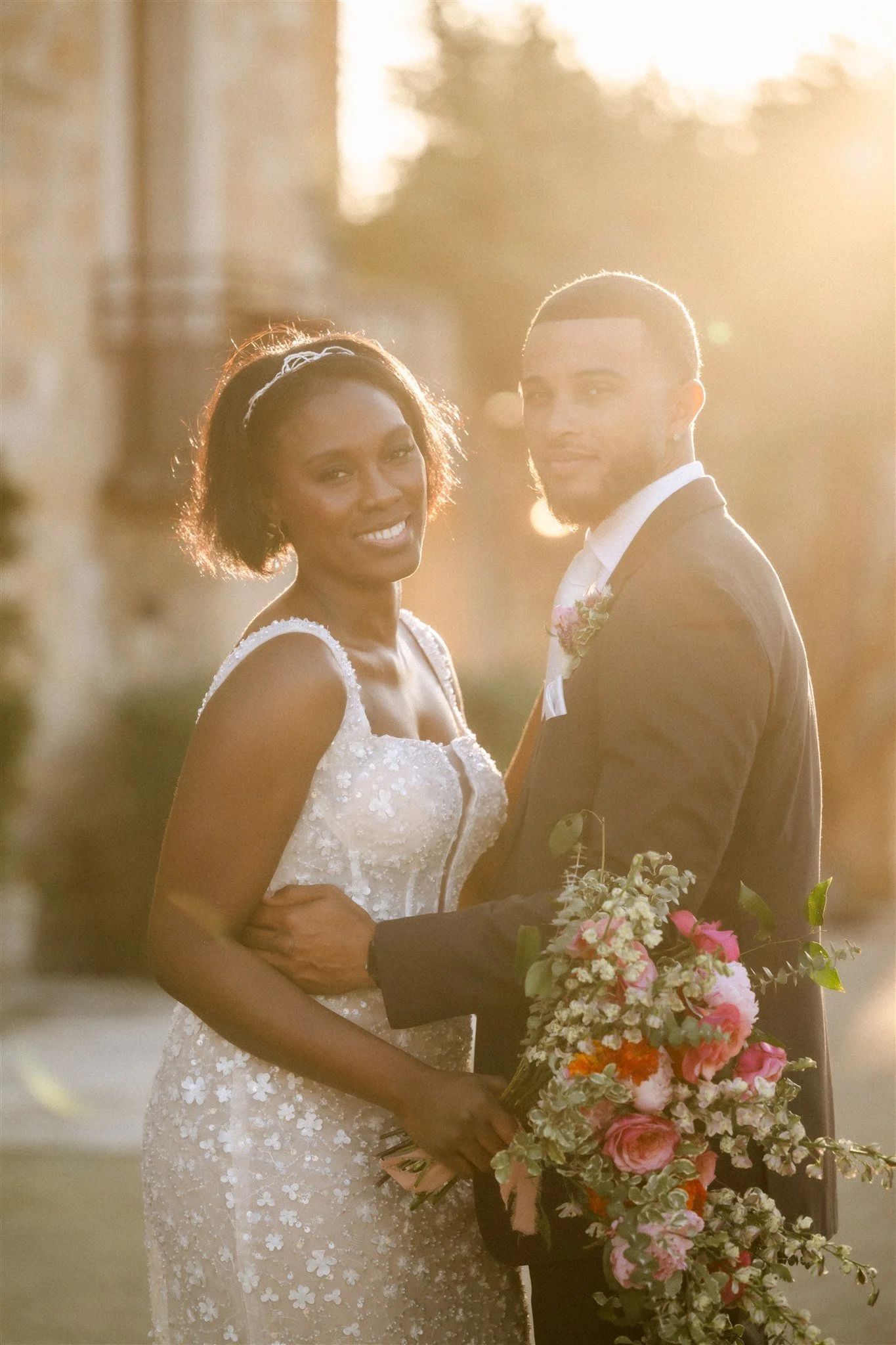 A bride and groom stand closely together outdoors during sunset, with the bride holding a bouquet of pink and white flowers.