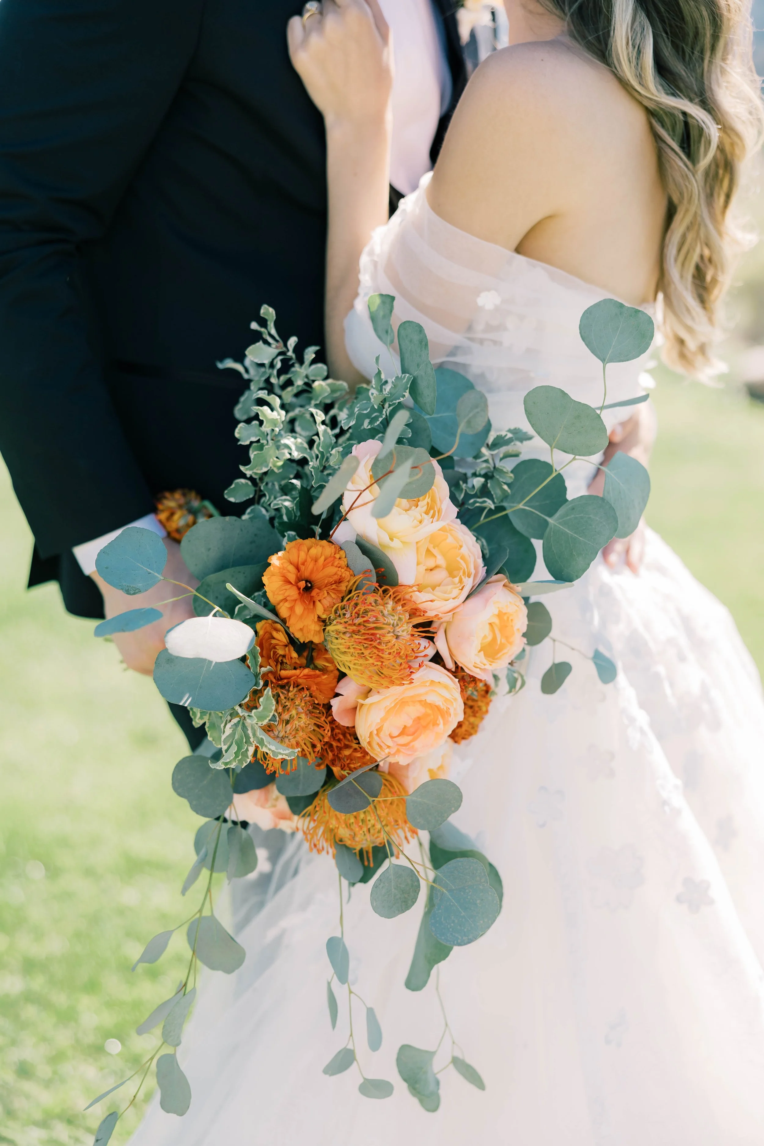 A bride holding a colorful bouquet of peach, orange, and yellow flowers with green eucalyptus leaves, standing outdoors.
