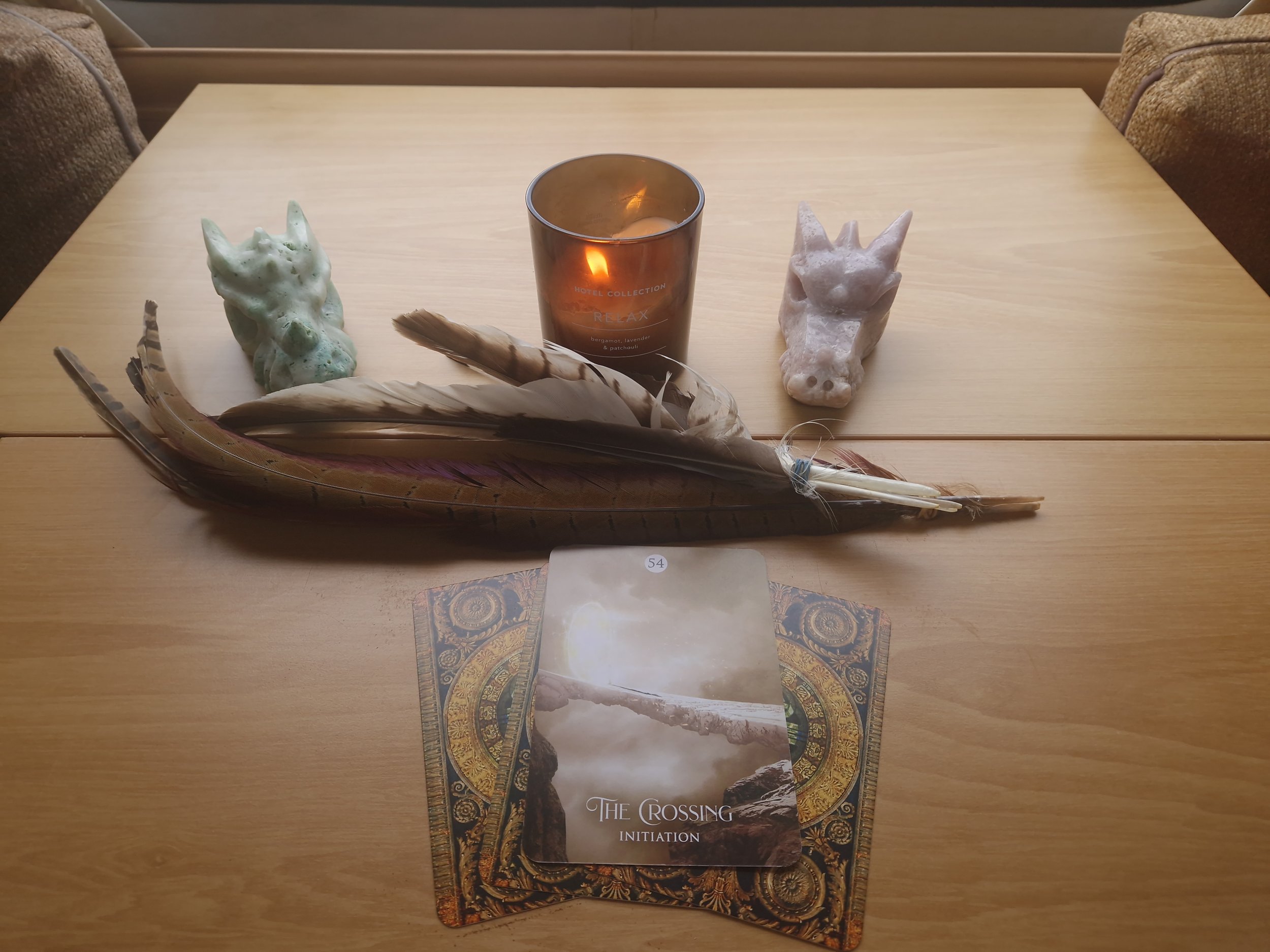 Brown table with a lite candle two dragon crystals one green and one pink in coloraturas. Feathers of a variety and 3 upside-down card from the shamans oracle deck and one card facing up with the words the crossing at the bottom of the card