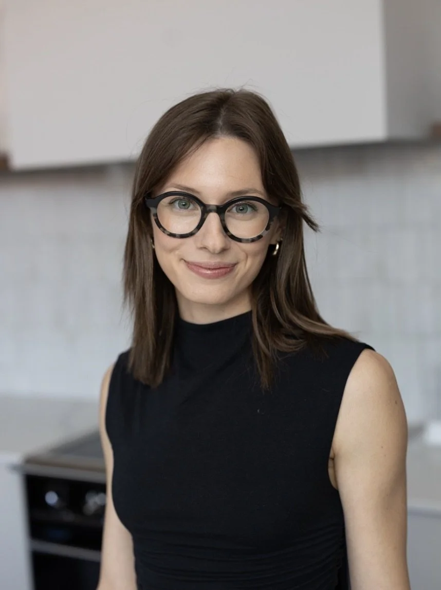 A woman with brown hair, glasses, and earrings wearing a sleeveless black top in a kitchen setting.