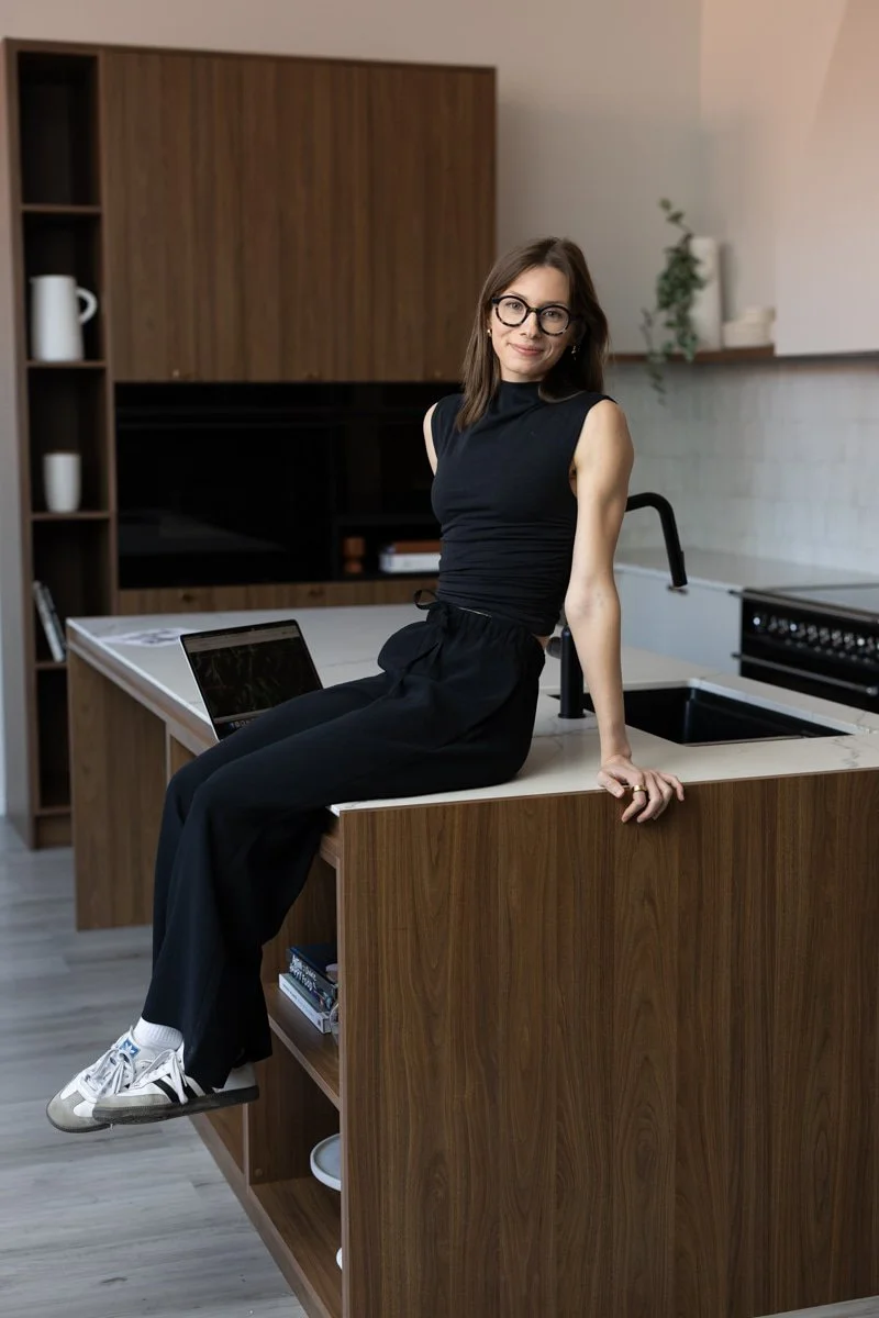 A woman with brown hair, glasses, and a black sleeveless top sitting on a kitchen island with a laptop, in a modern kitchen with wood cabinetry and a plant in the background.