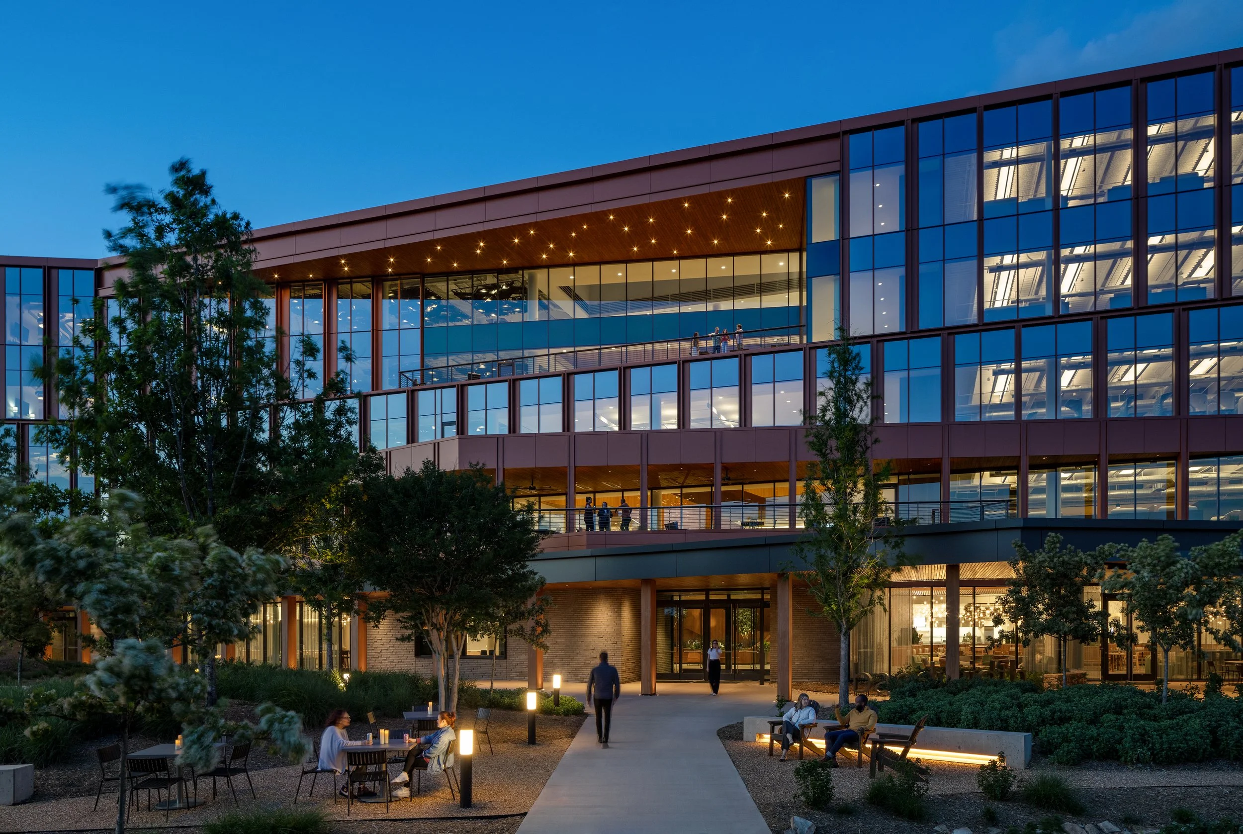 Modern office building with glass facade, illuminated at dusk, with people sitting and walking outside.