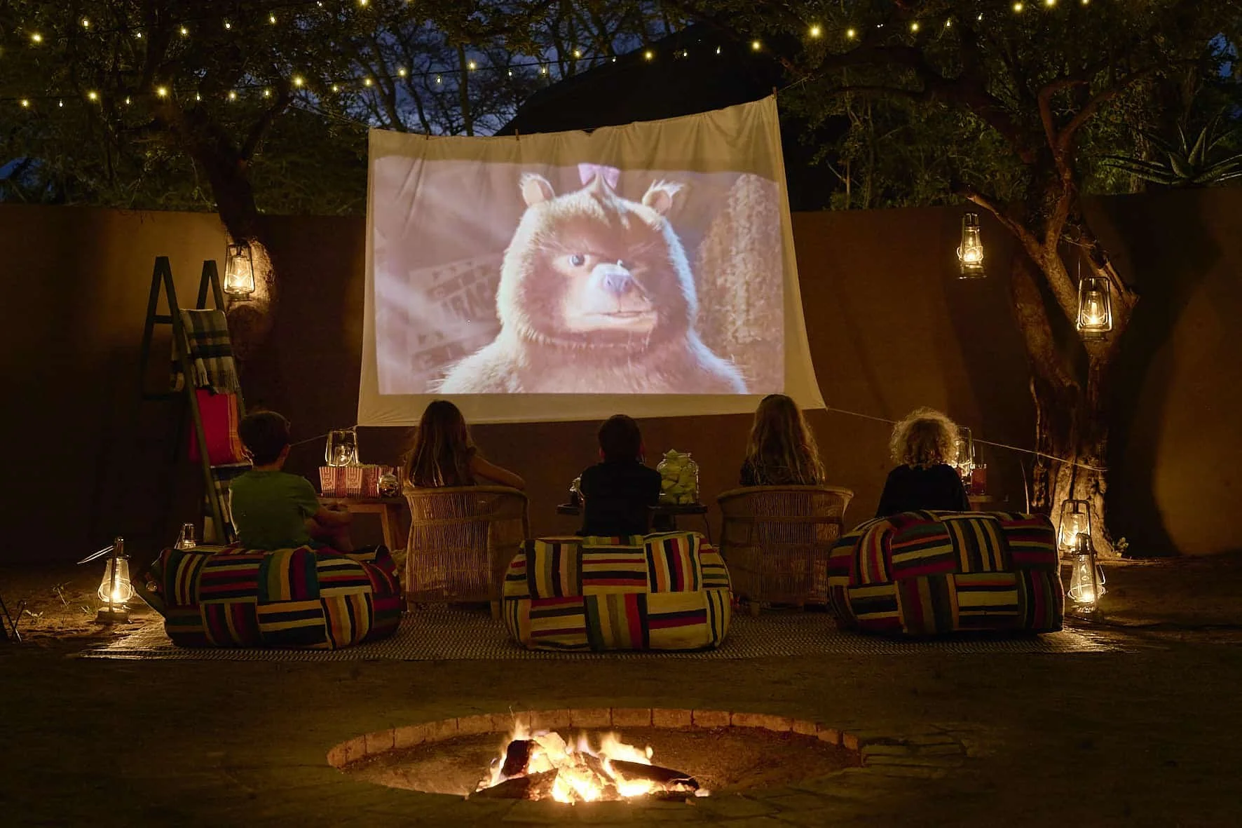 People sitting outdoors around a fire pit watching a movie projected on a screen at night, with string lights and lanterns hanging from trees.