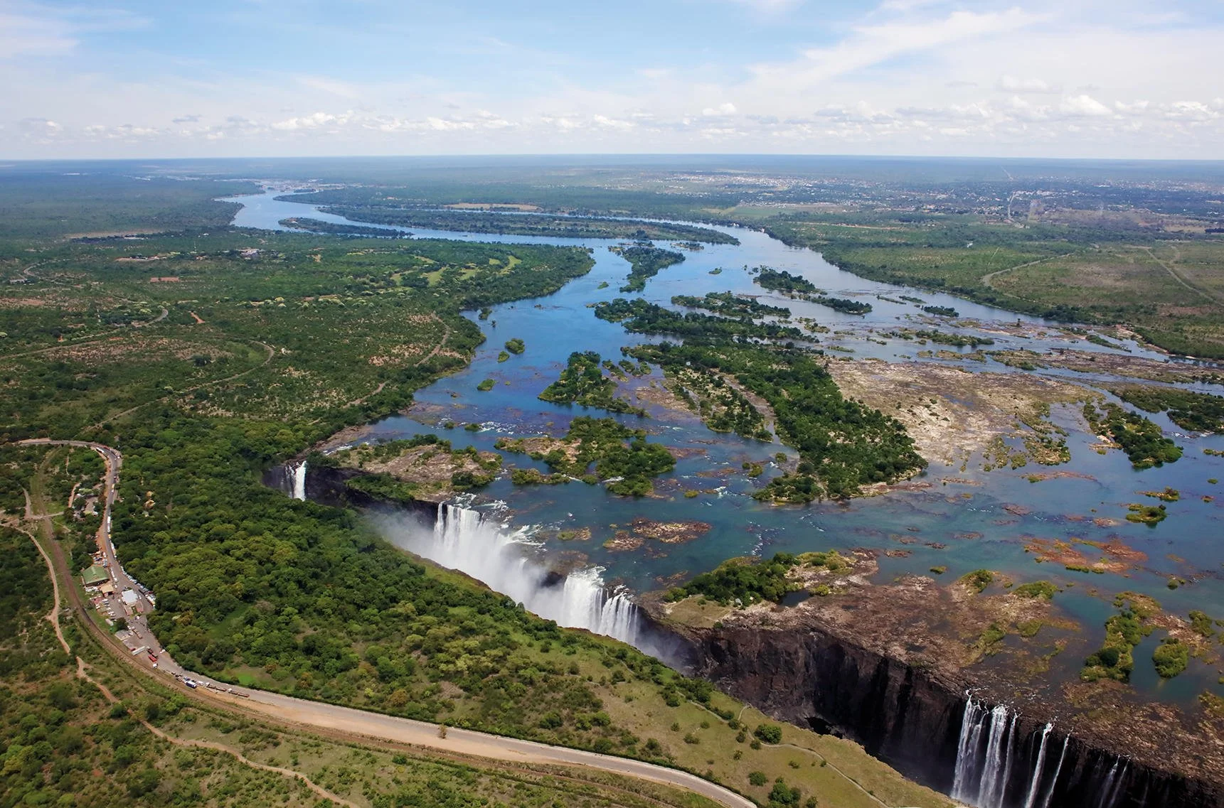 Aerial view of waterfalls in a lush, green landscape with a river, islands, and surrounding terrain, under a partly cloudy sky.
