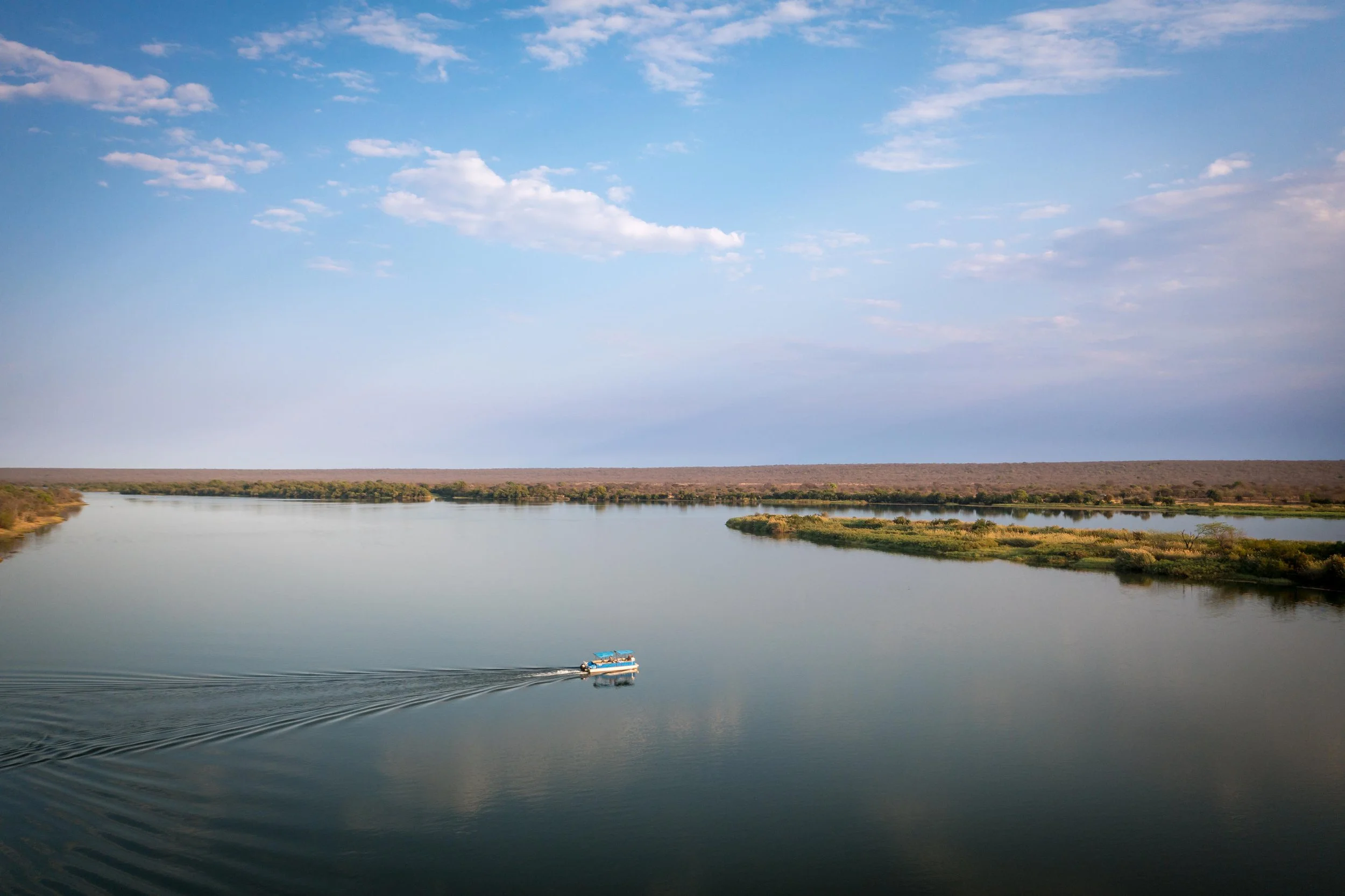A boat with a blue canopy sailing on a wide river under a partly cloudy sky, with greenery along the riverbanks and a flat horizon in the distance.