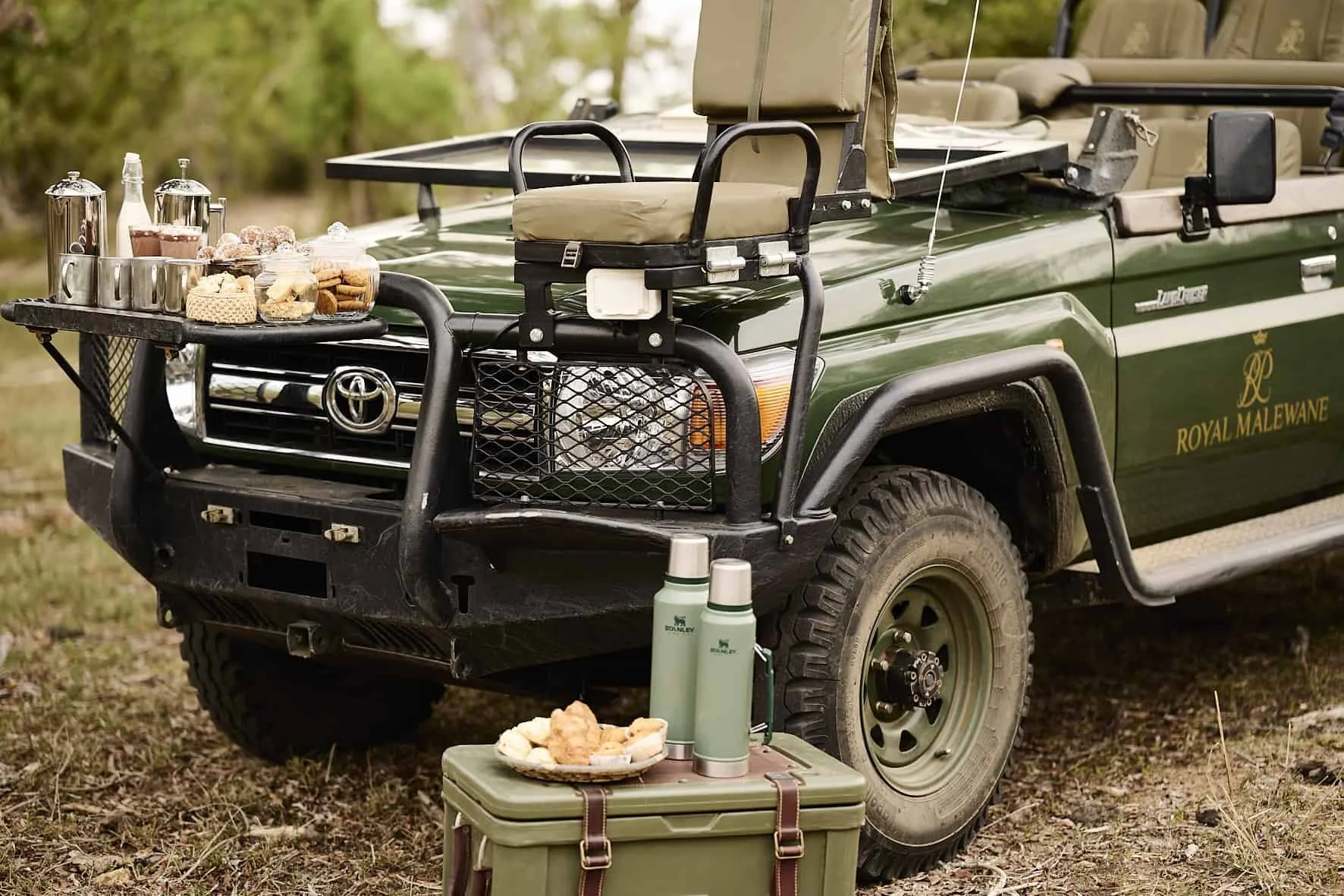 A green Toyota Land Cruiser with a front rack carrying coffee, tea, cookies, and snacks on a biscuit tray. There are two green thermoses and a plate with food on a green insulated cooler in front of the vehicle. The vehicle has a logo 'Royal Malewane' on the side and is parked on grass in a natural outdoor setting.