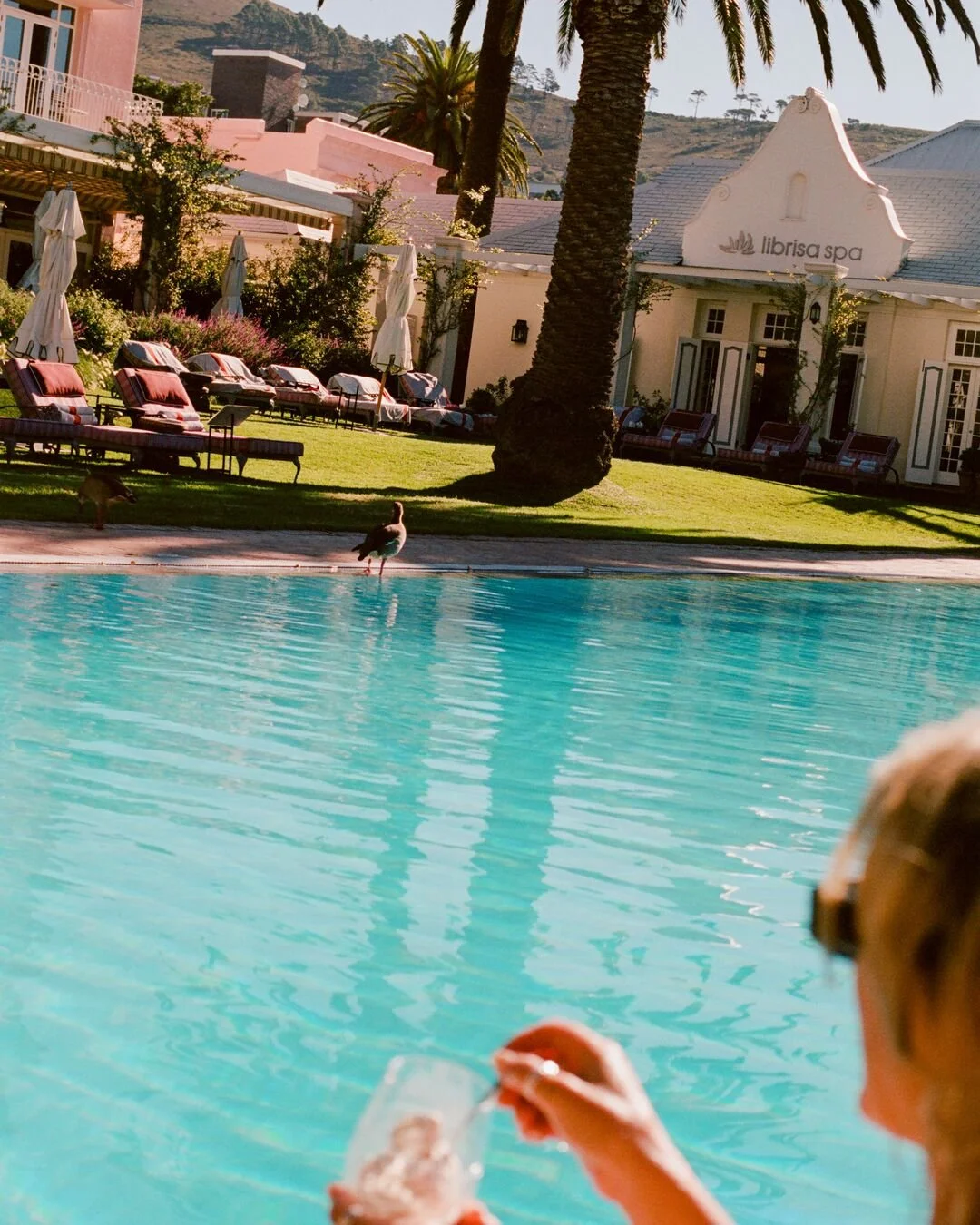 A person holding a drink by a swimming pool with palm trees, lounge chairs, umbrellas, and a spa building in the background.