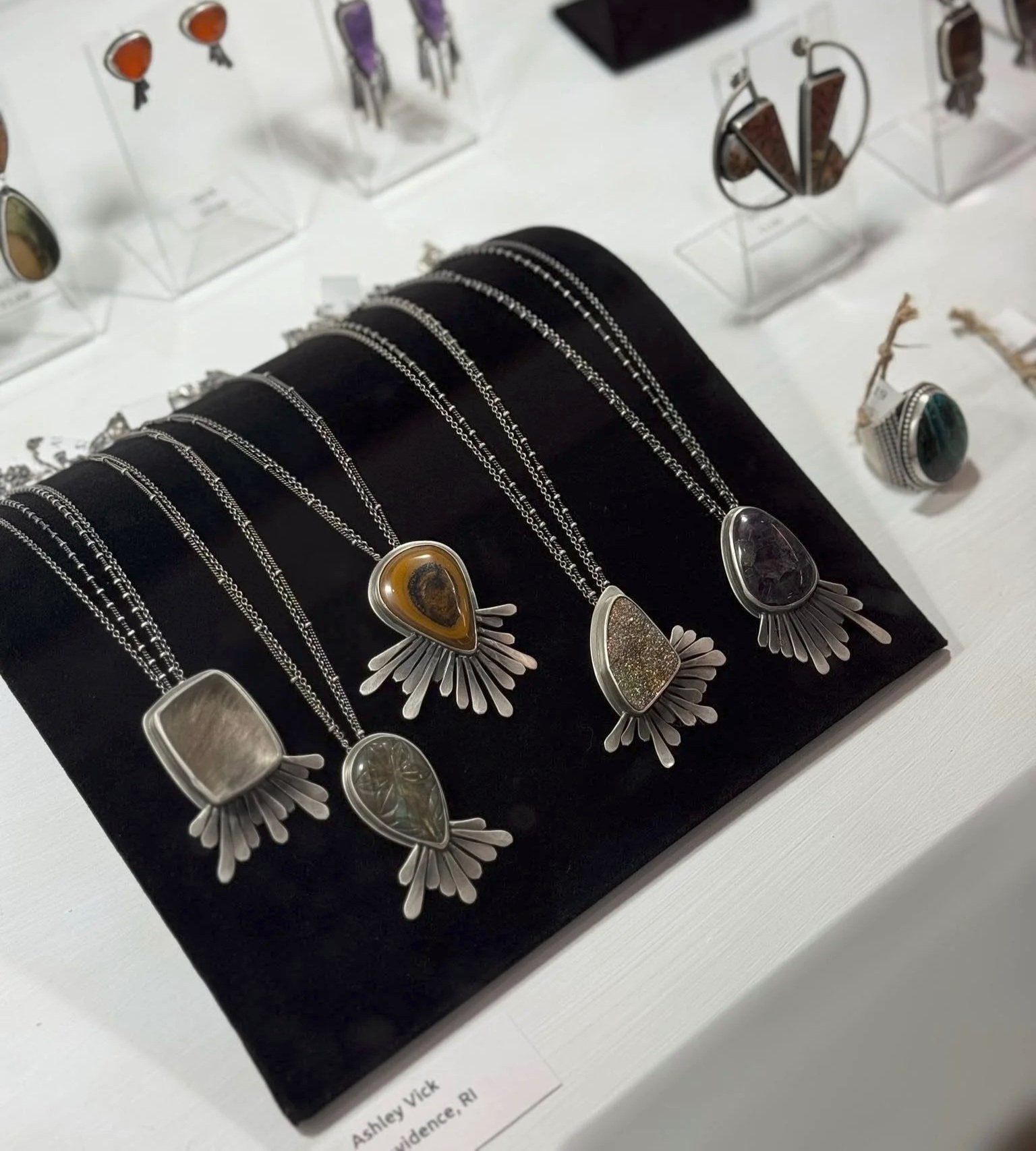 Display of silver jewelry necklaces and rings with large gemstone pendants and intricate designs on a black stand, with other jewelry pieces in the background.