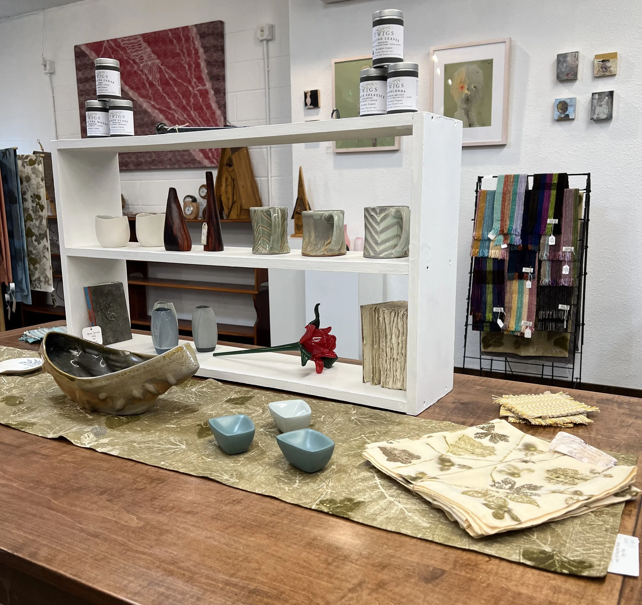 A retail store display with decorative pottery by Joy Tanner on a white stand, colorful fabric scarves  by Edwina Bringle on a black rack, and framed artwork by Connie Karleta-Sales on the wall in the background.
