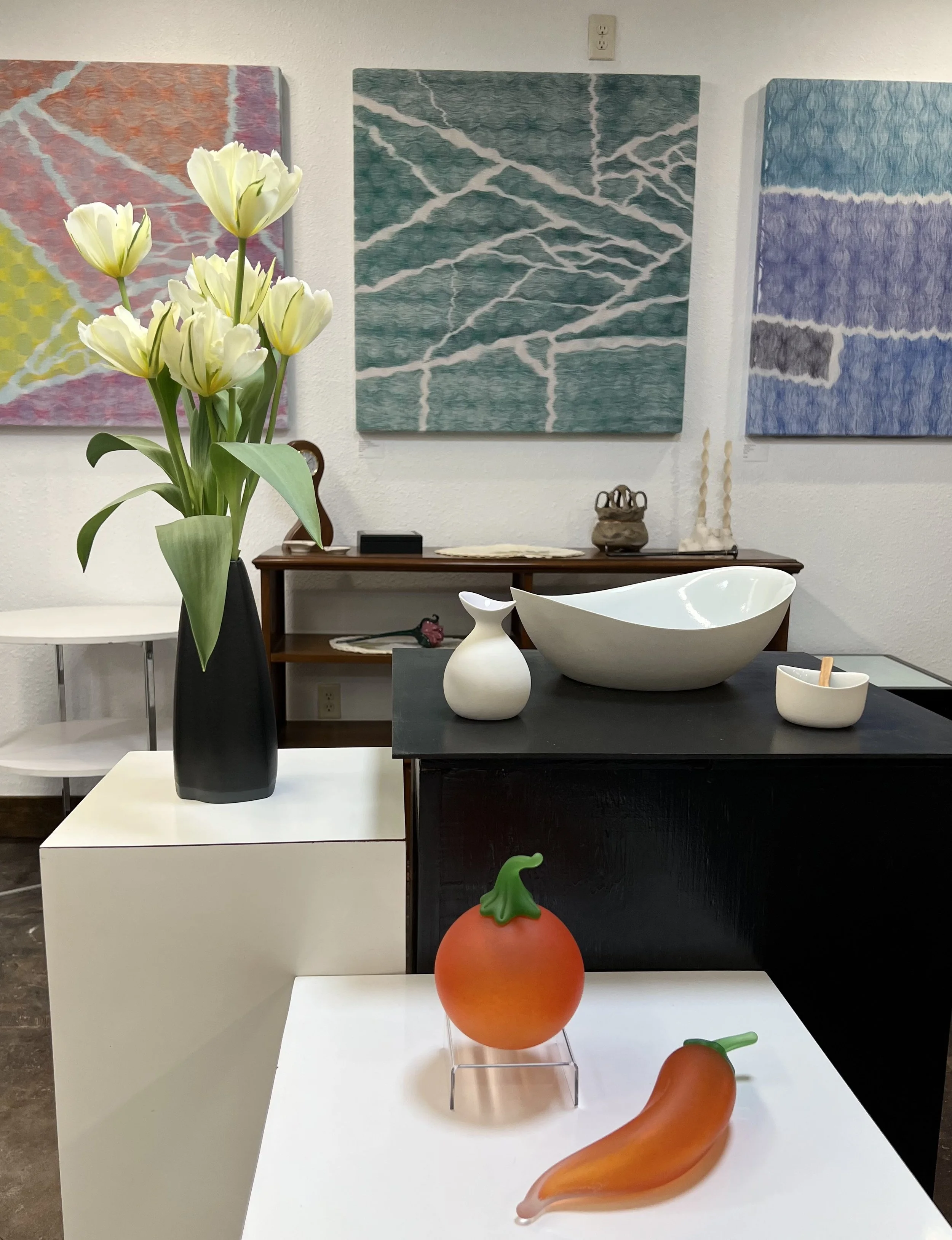 Colorful abstract weavings on a white wall behind a display of modern decorative objects including a black Stormie Burns vase with white tulips, a white pitcher, a white bowl, glass pepper and tomato by Rob Levin, a set of white candles, and a small 