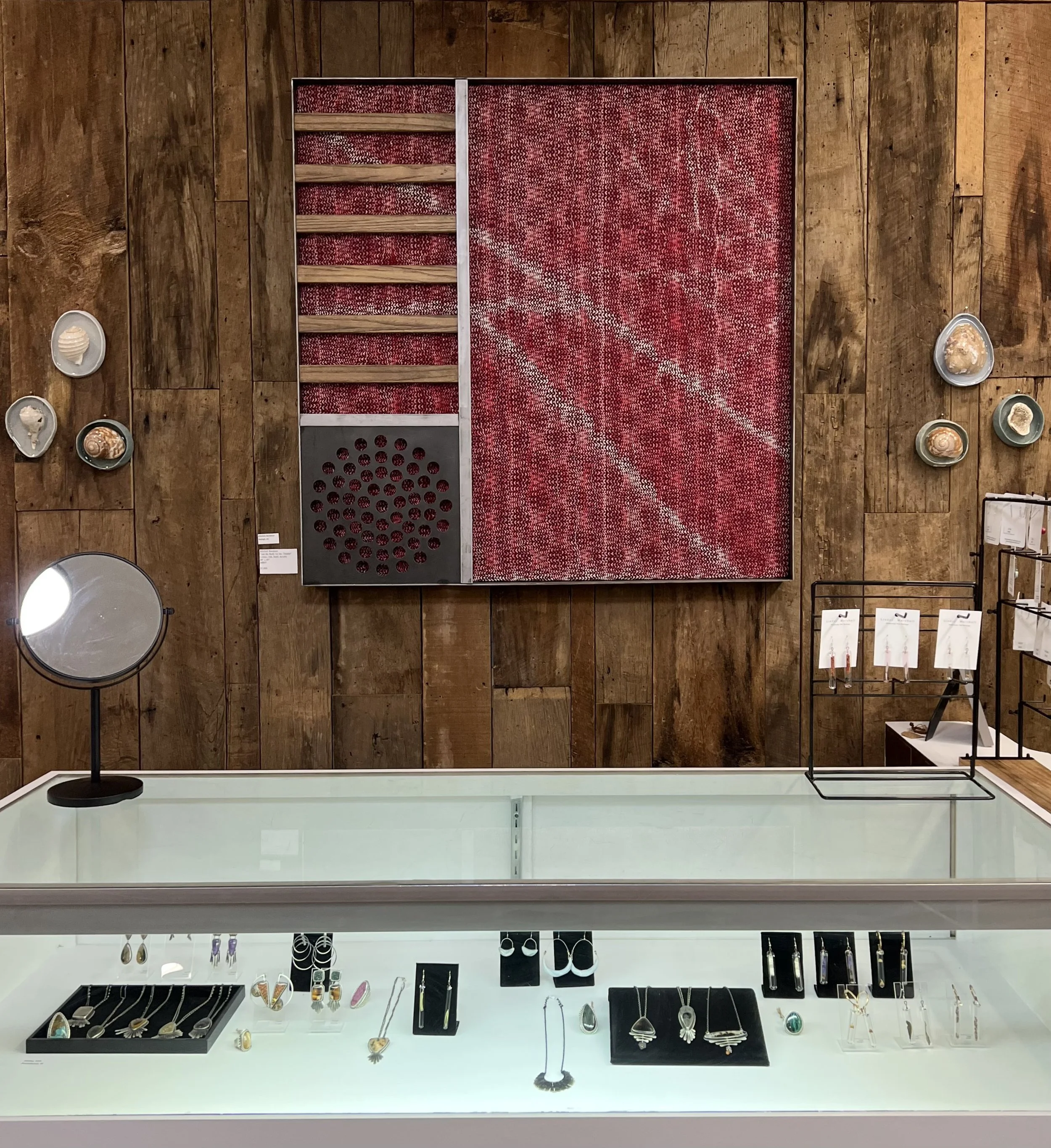 Display of jewelry including rings, earrings, necklaces, and bracelets on black and white stands. With a large weaving by Mitchell Burleson above.