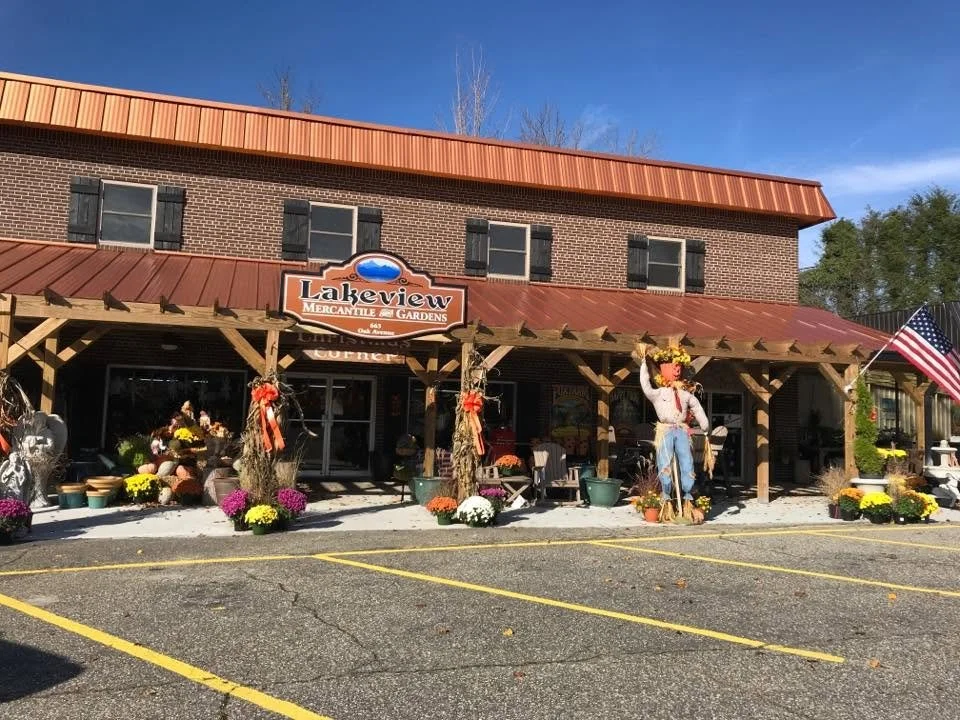 The exterior of a brick building decorated with fall harvest decorations including pumpkins, flowers, and scarecrows, with outdoor seating, a parking lot, and an American flag on the right side.