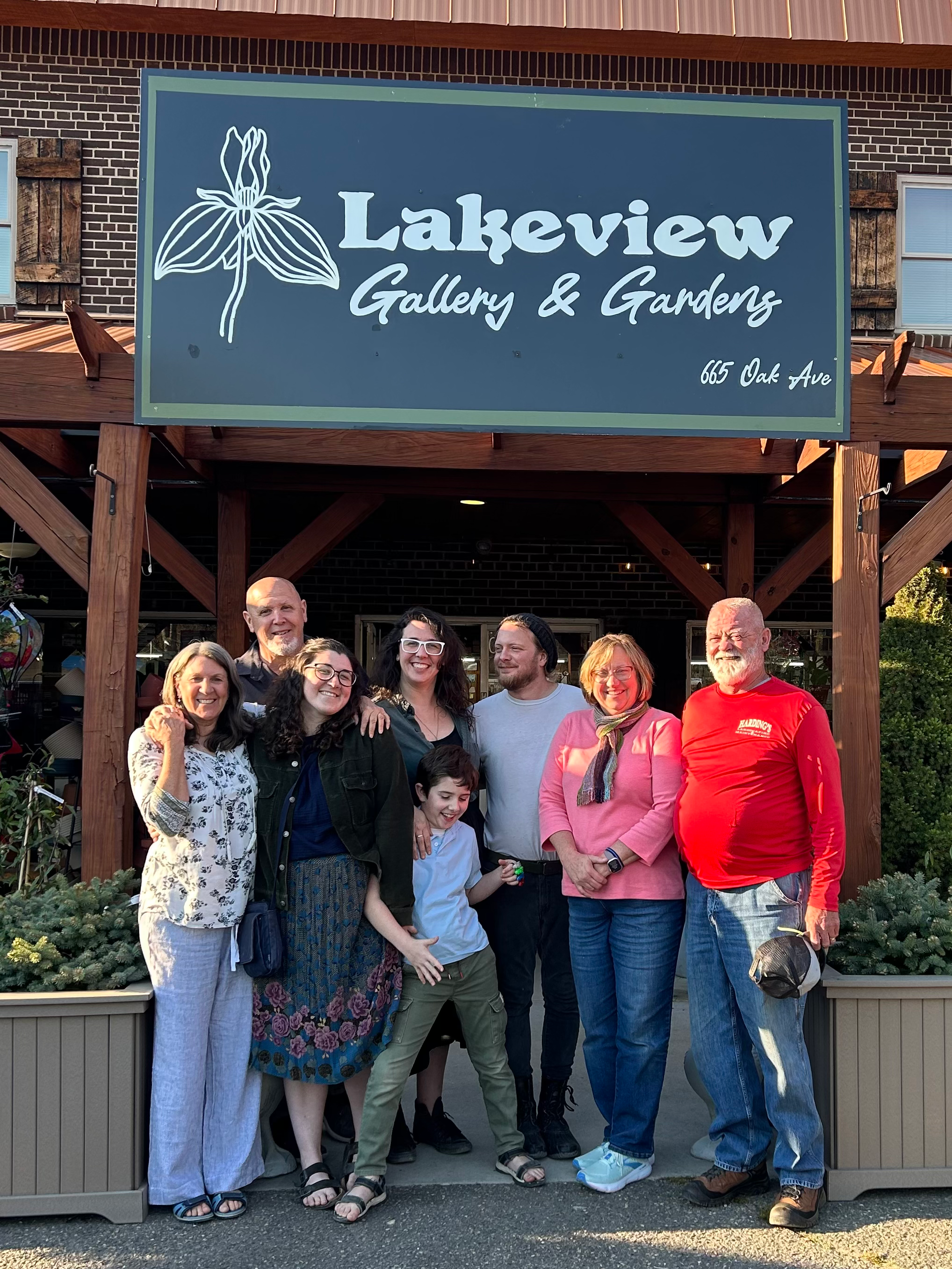 A group of ten  people, including current and former owners of Lakeview, posing together outside in front of a sign that reads 'Lakeview Gallery & Gardens' on Oak Avenue, with some plants and greenery at the front.