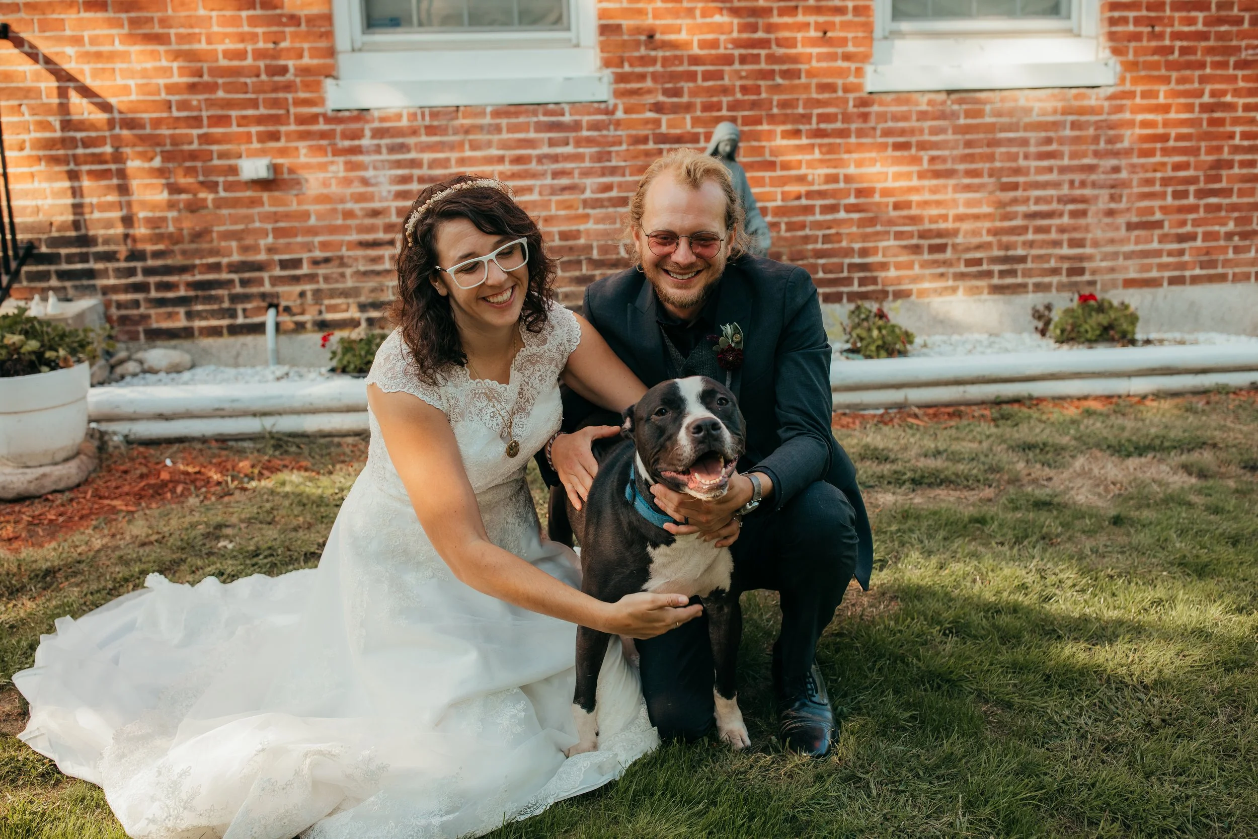 Lakeview Gallery and Gardens owners, Faith and Ryan Byrd, holding a happy black and white dog during an outdoor wedding celebration in front of a brick wall.