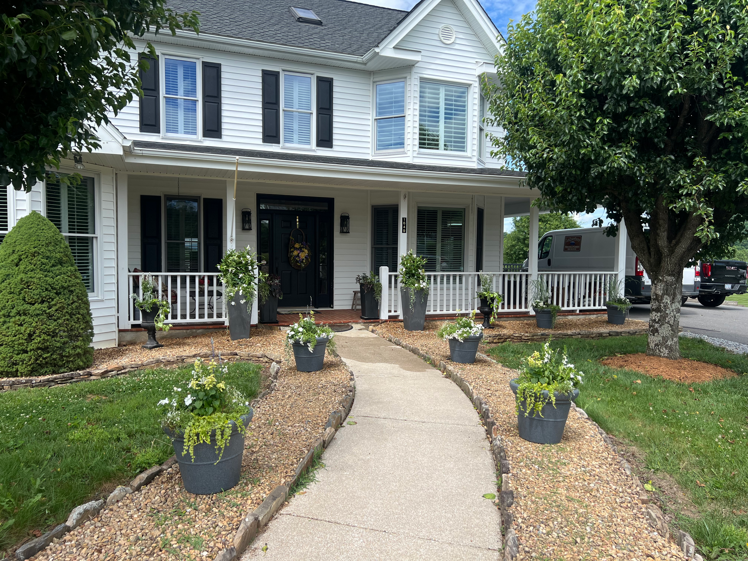 Front yard of a white house with black shutters, a covered porch, and potted plants lining a curved walkway, flanked by trees and greenery.