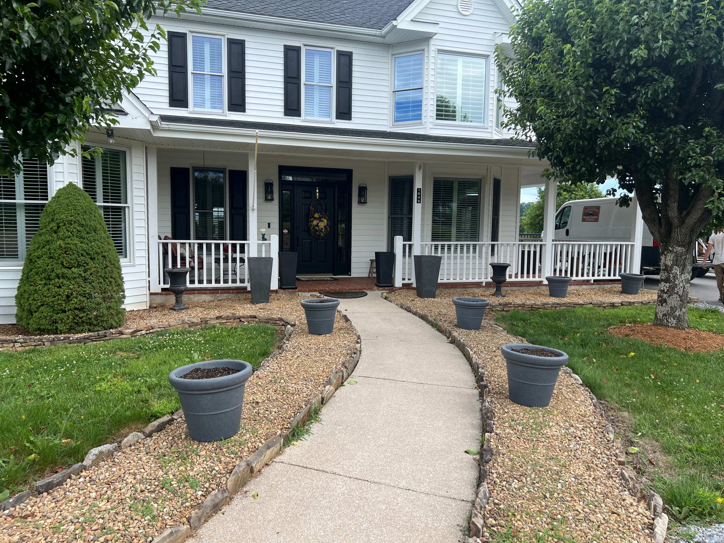 Front view of a white two-story house with black shutters, a black front door with a wreath, a white porch railing, a sidewalk leading to the porch, before planters are potted along the walkway, and trees on both sides.