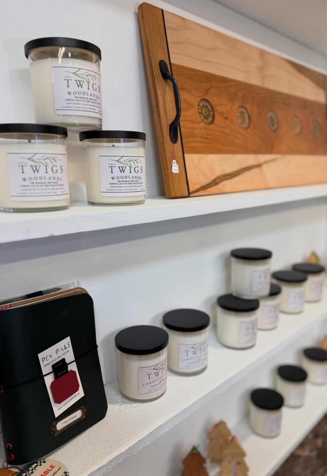 Display of scented candles on white shelves, with a wooden cutting board and small wooden christmas ornaments in the background.