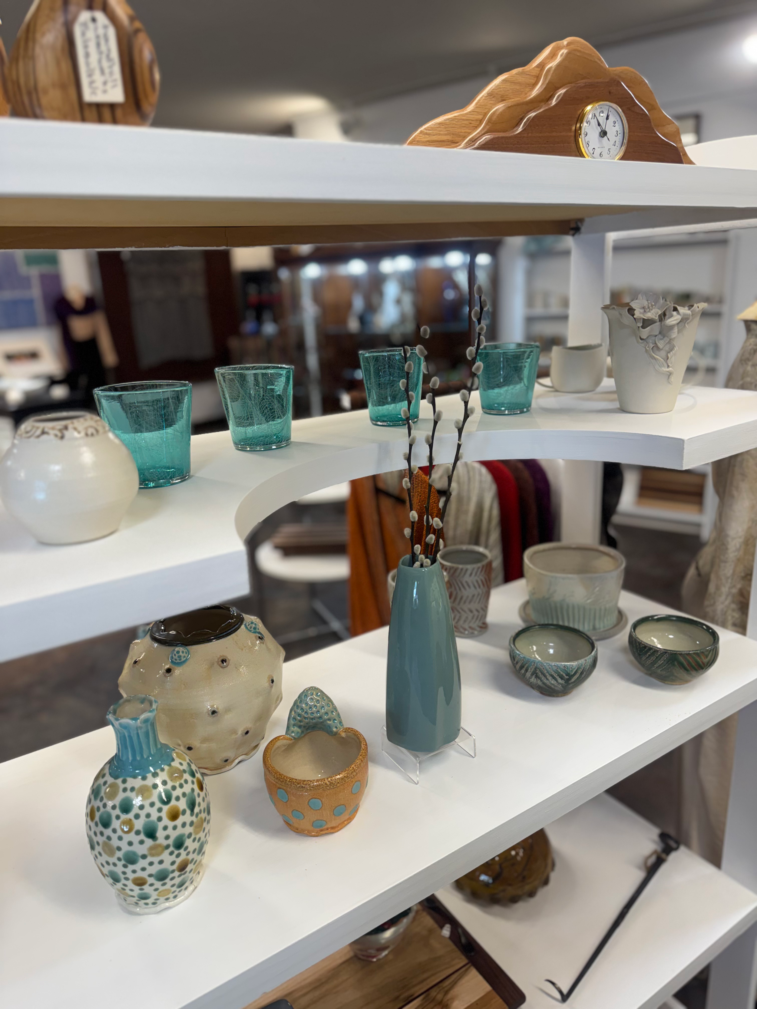 Display of decorative ceramic vases and bowls by Ronan Kyle Peterson and Marianne Cicala predominately on white shelves in a store.