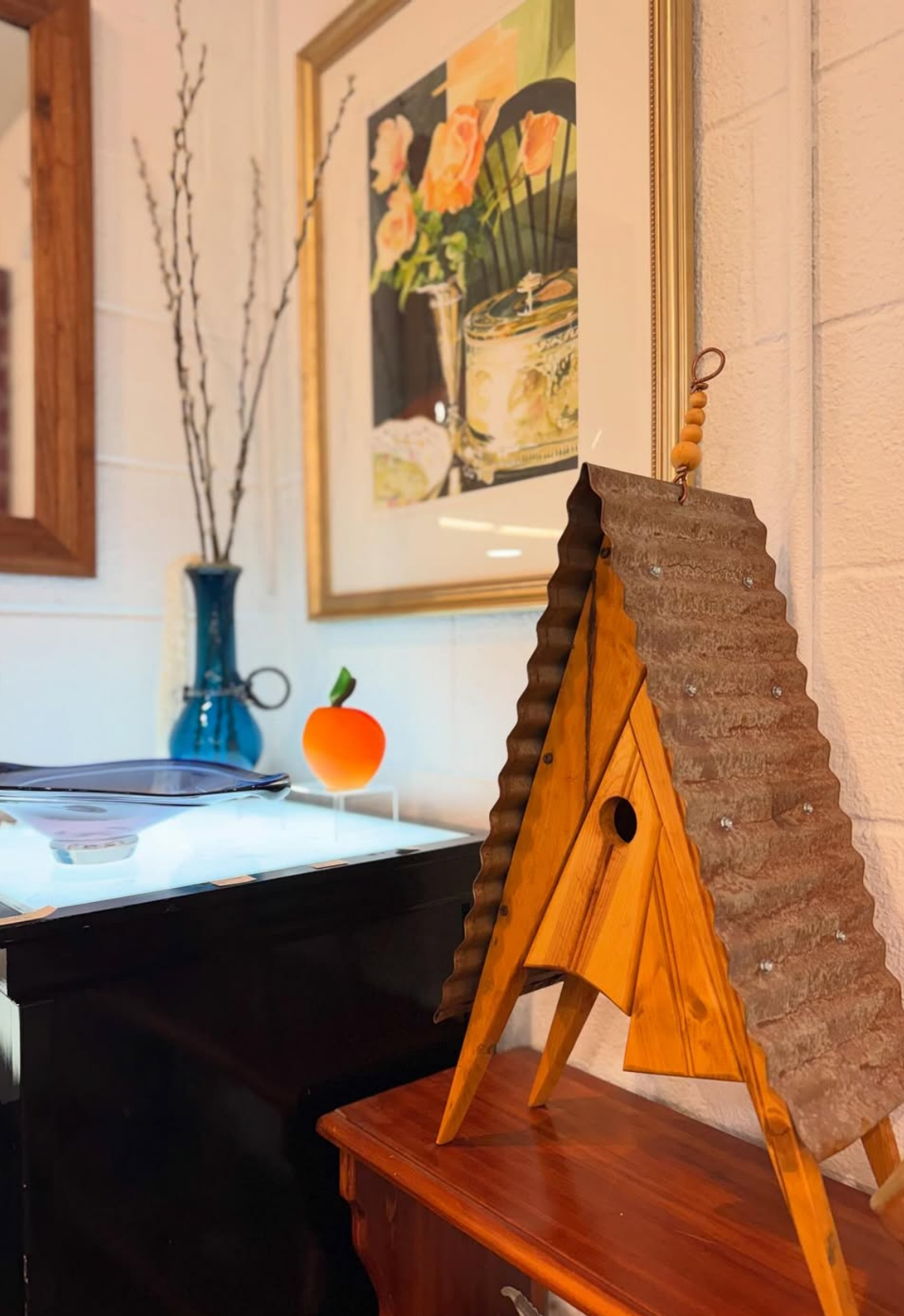 Decorative wooden birdhouse by Lynn Alinger with corrugated roof placed on a small wooden table, with framed floral paintings on the wall.