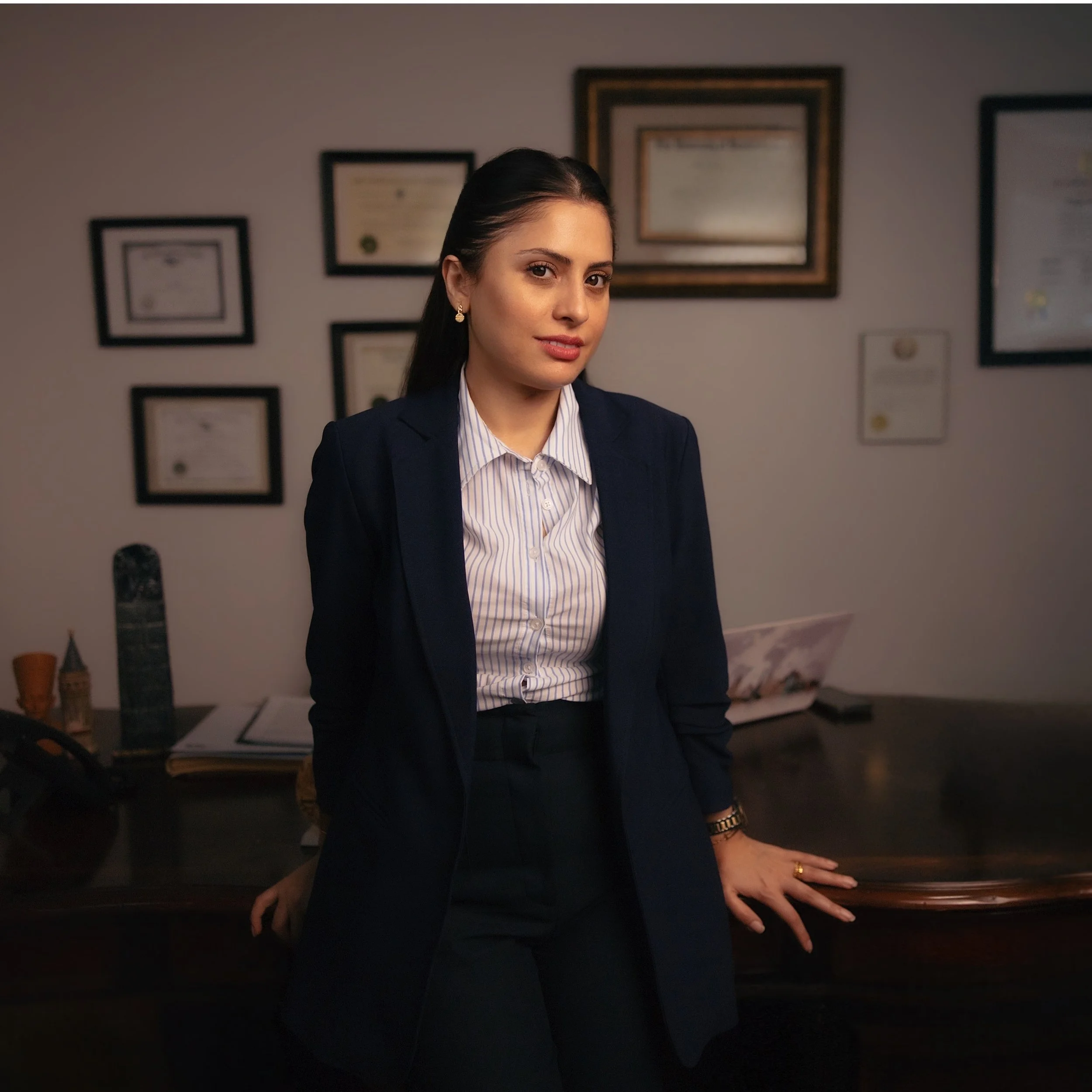 A woman with dark hair and light skin wearing business attire, including a navy blazer and striped shirt, standing with one hand resting on a dark surface in a room with framed certificates or diplomas on the wall in the background.