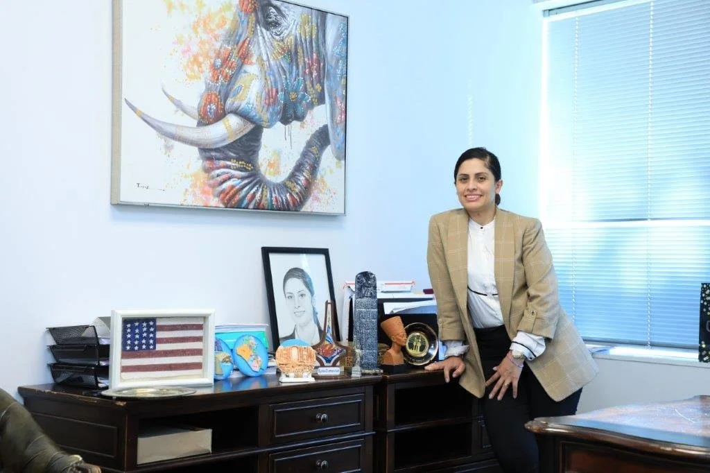A woman in beige blazer standing in an office with framed photos, a painting of an elephant, and office supplies on a dark wooden cabinet, with blinds and a window behind her.